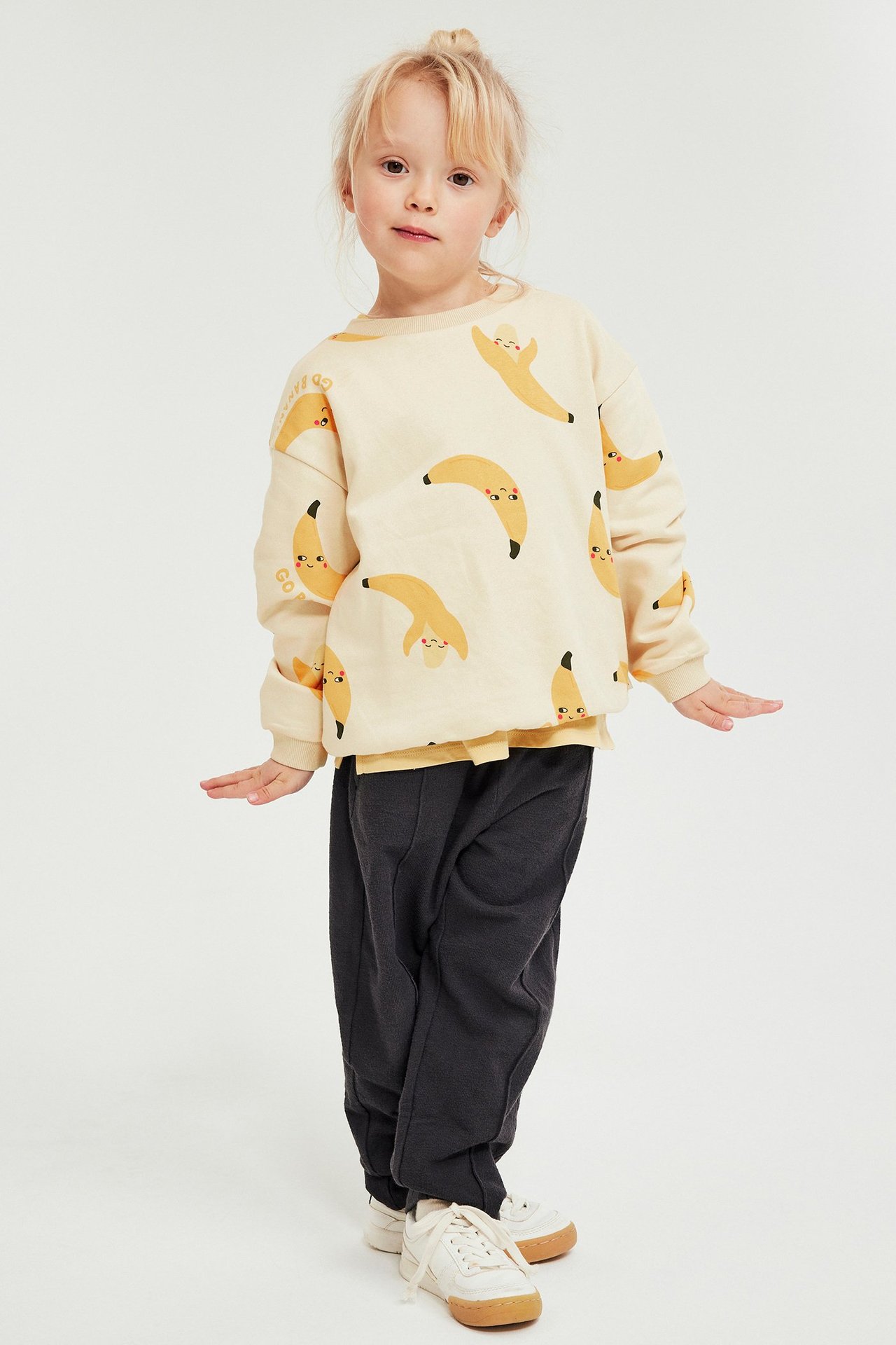 Edestäpäin: lapsi beigessä banaanikuvioisessa collegepaidassa, tummanharmaissa housuissa ja valkoisissa tennareissa.
