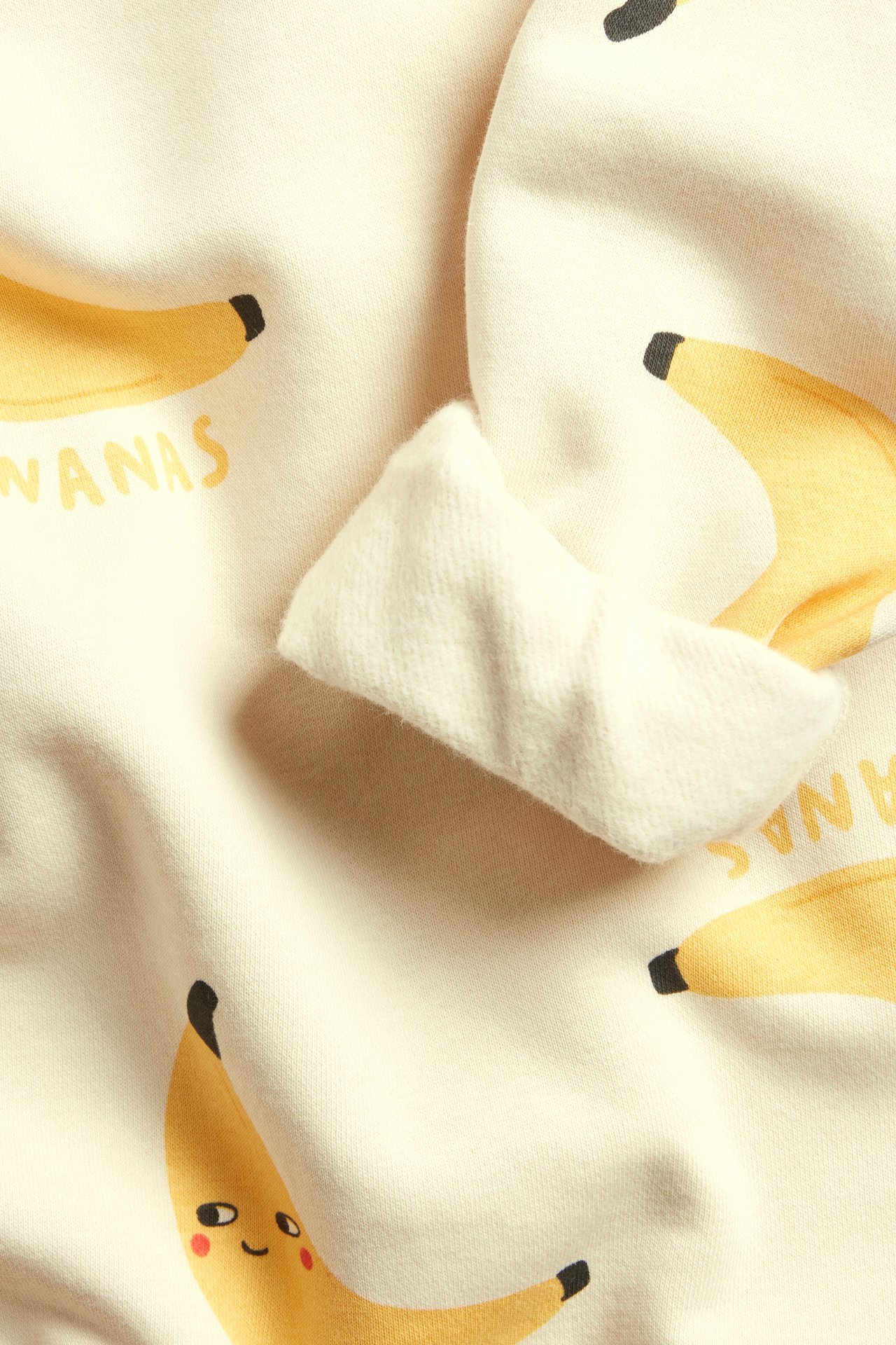 Nærbilde av en beige sweatshirt i bomullsblanding med gult banantrykk og synlig ribbet mansjett.