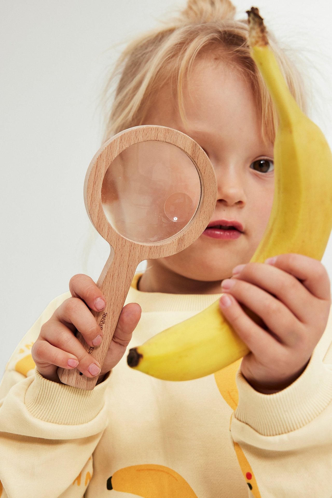 Lähikuva edestä lapsesta beigessä banaaniprinttihupparissa, joka pitää puista suurennuslasia ja banaania.