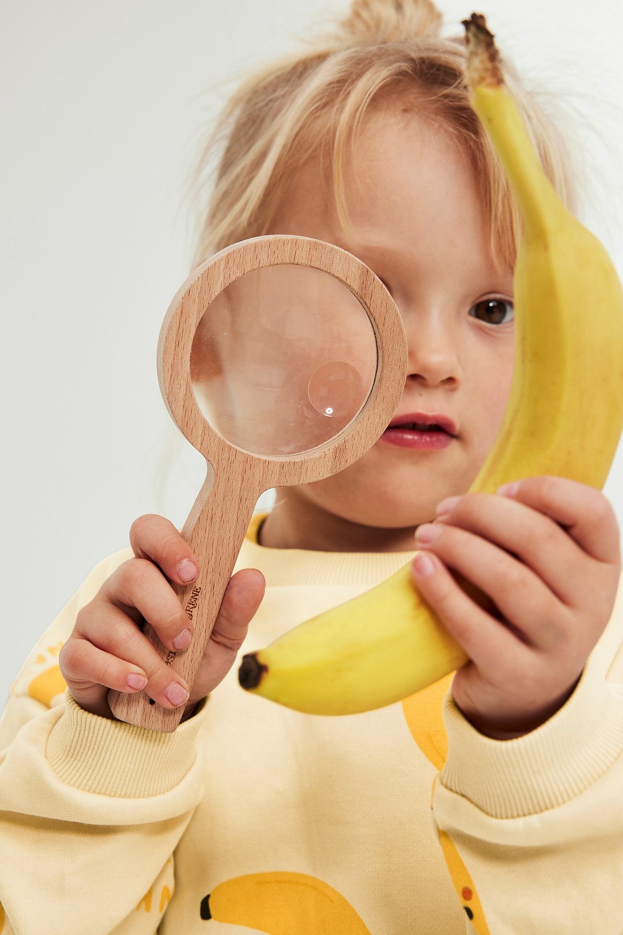Nærbilde forfra av et barn i en lys gul sweatshirt med bananmønster, som holder et forstørrelsesglass og en banan.