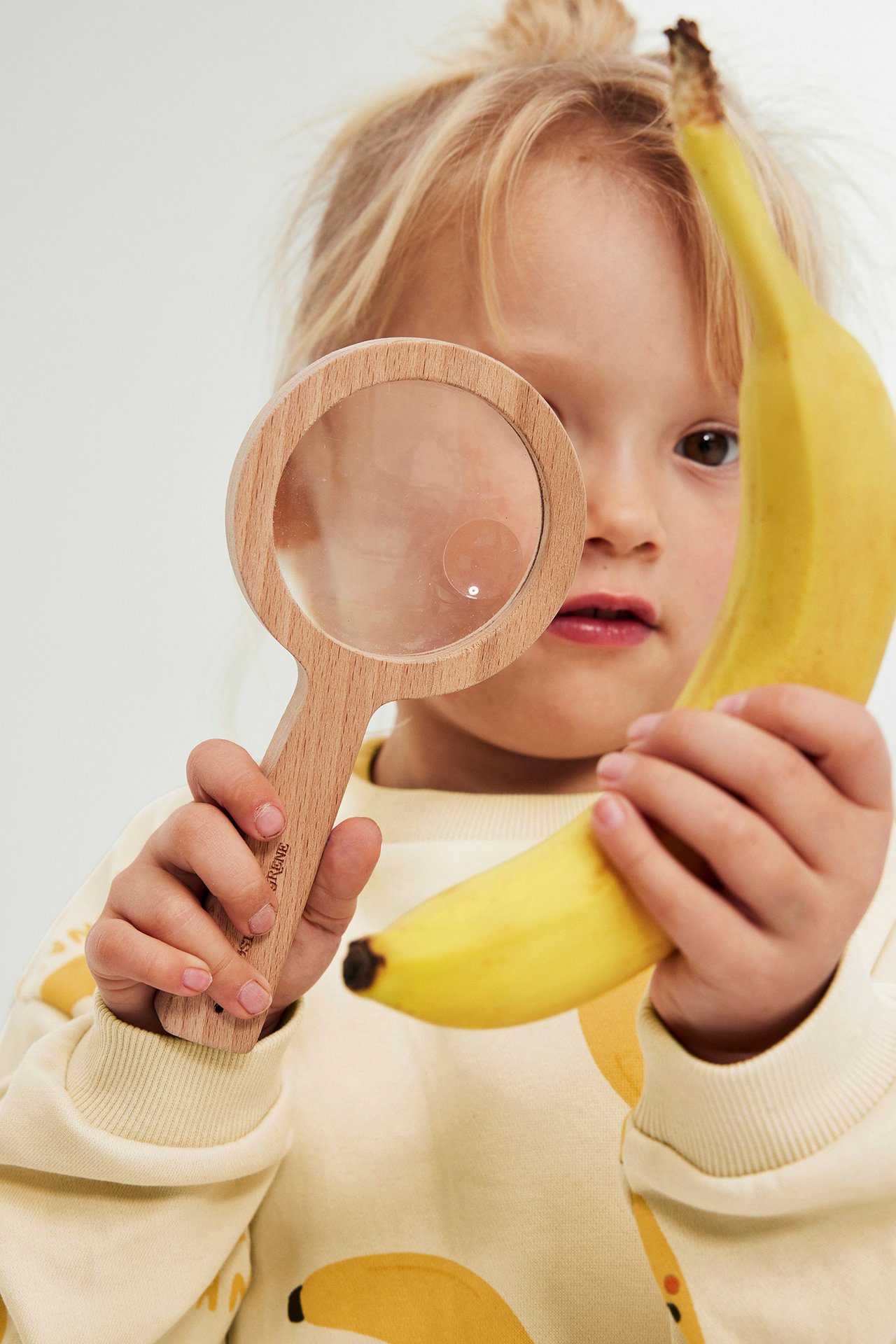 Zbliżenie dziecka w beżowej bluzie z nadrukiem bananów, trzymającego lupę i banana.
