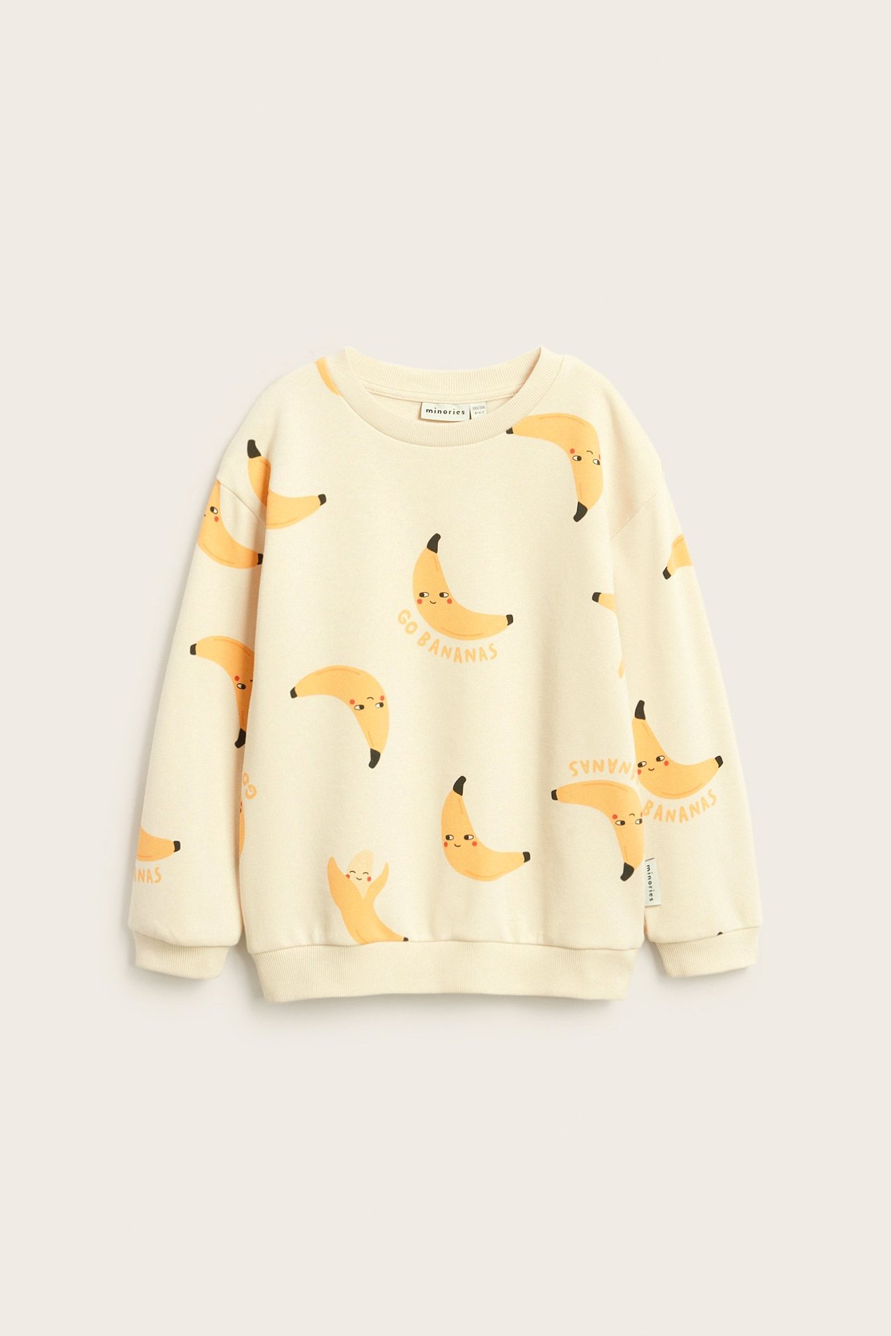Forfra, en beige barnesweatshirt med gjentatt mønster av smilende bananer og ribbestrikkede mansjetter.