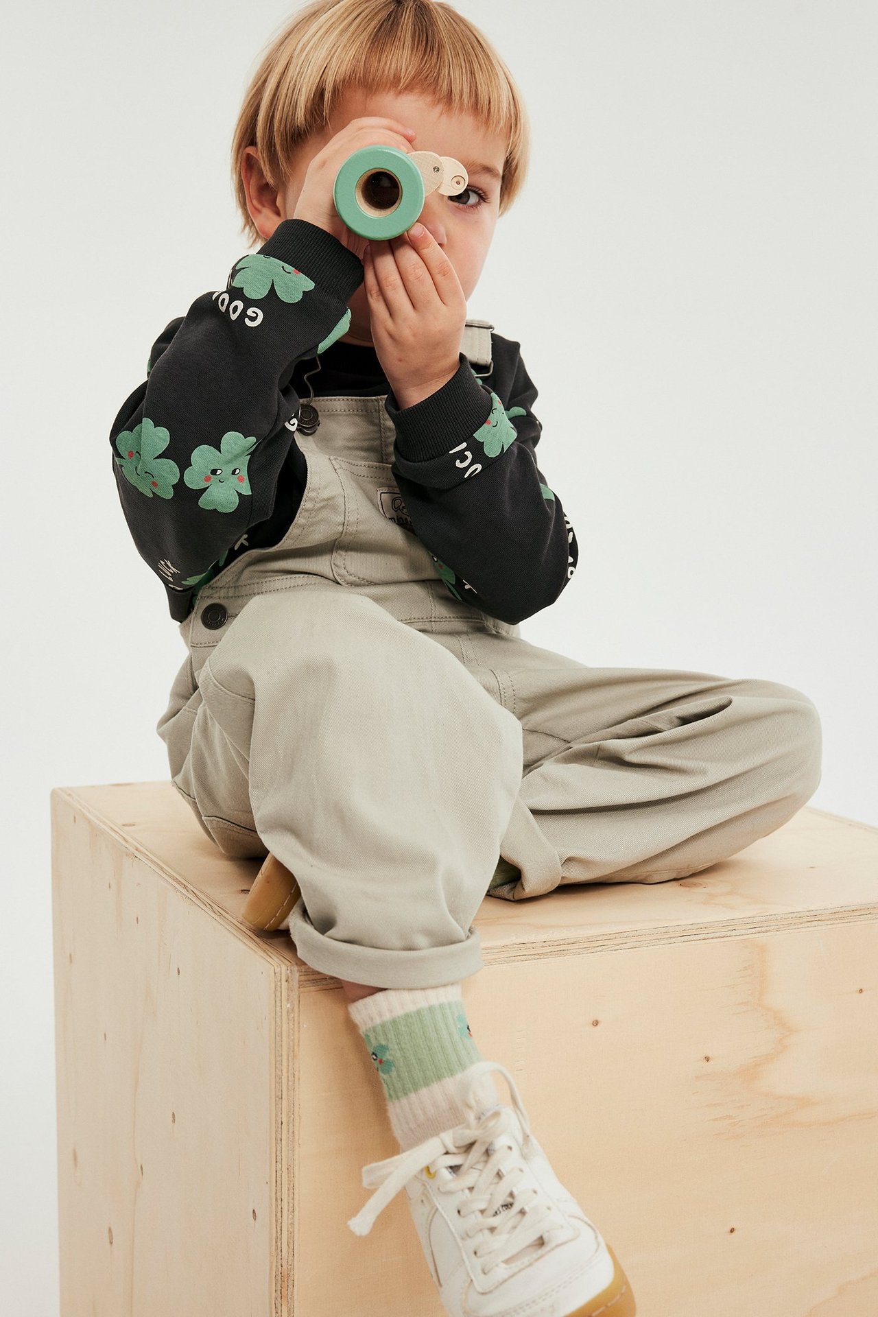 Edestä: lapsi mustassa apilacollagepaidassa, beigeissä haalareissa, valkoisissa tennareissa, pitää vihreää lelua.