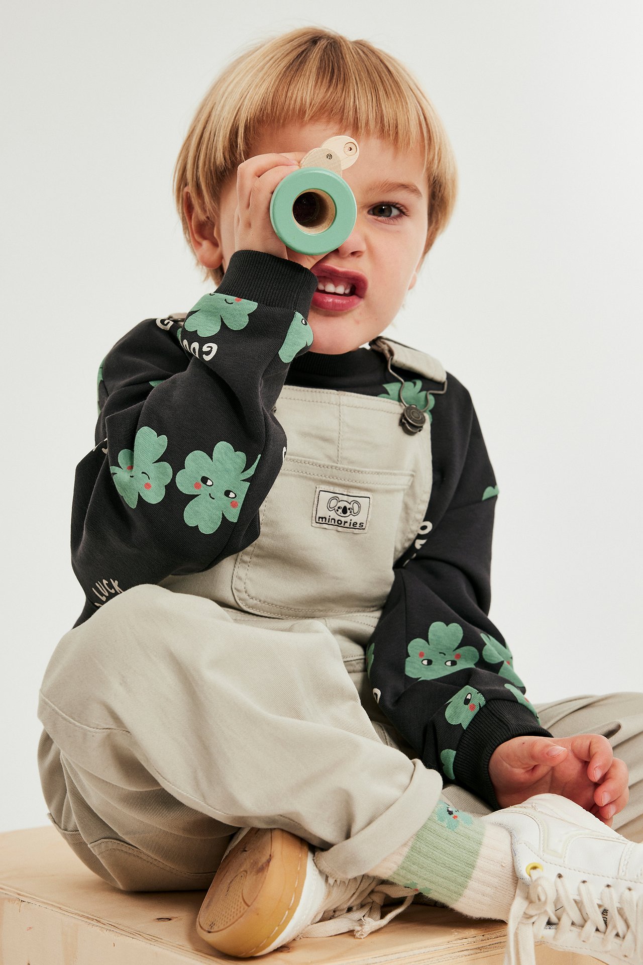 Lapsi edestä mustassa kuviollisessa collegepaidassa vihreillä apilanlehdillä vaaleanbeigen haalarin päällä.