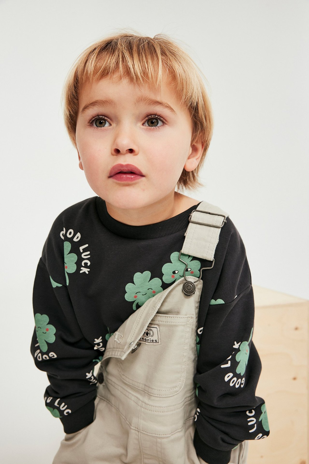 Edestä, lapsi mustassa collegepaidassa vihreällä apilakuviolla, vaaleanbeigien haalareiden päällä.