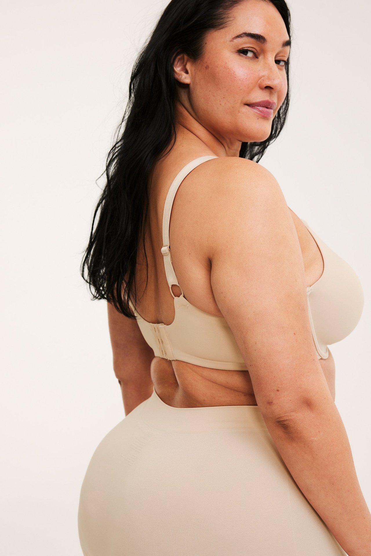 Sidebilde av en kvinne i en lys beige minimizer-bh med spile og matchende sømløse underbukser.