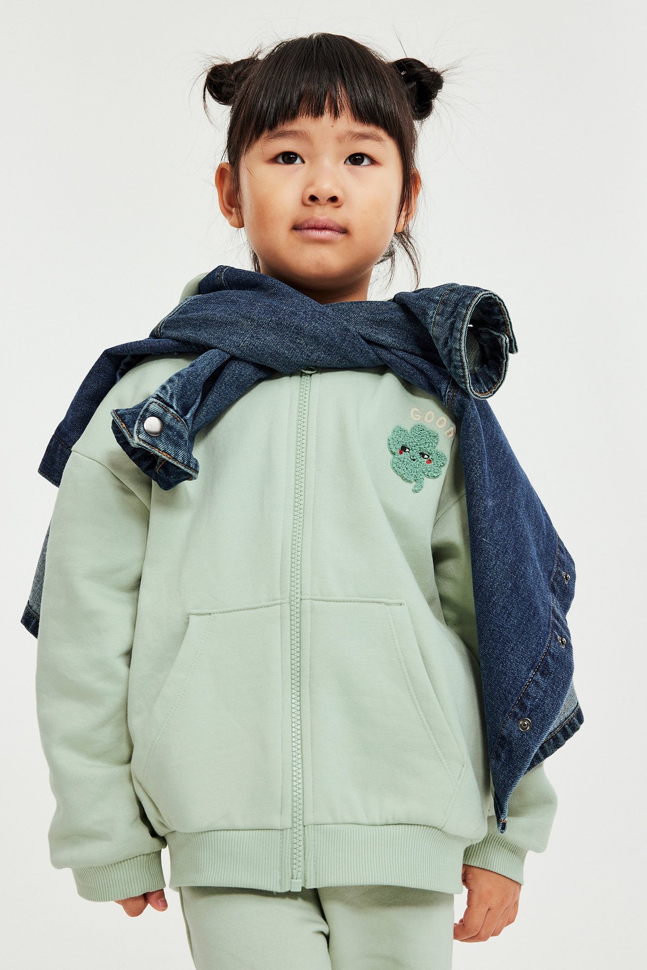 Widok z przodu dziecka w jasnozielonej bluzie z zamkiem, naszywką koniczyny, z kurtką dżinsową na ramionach.