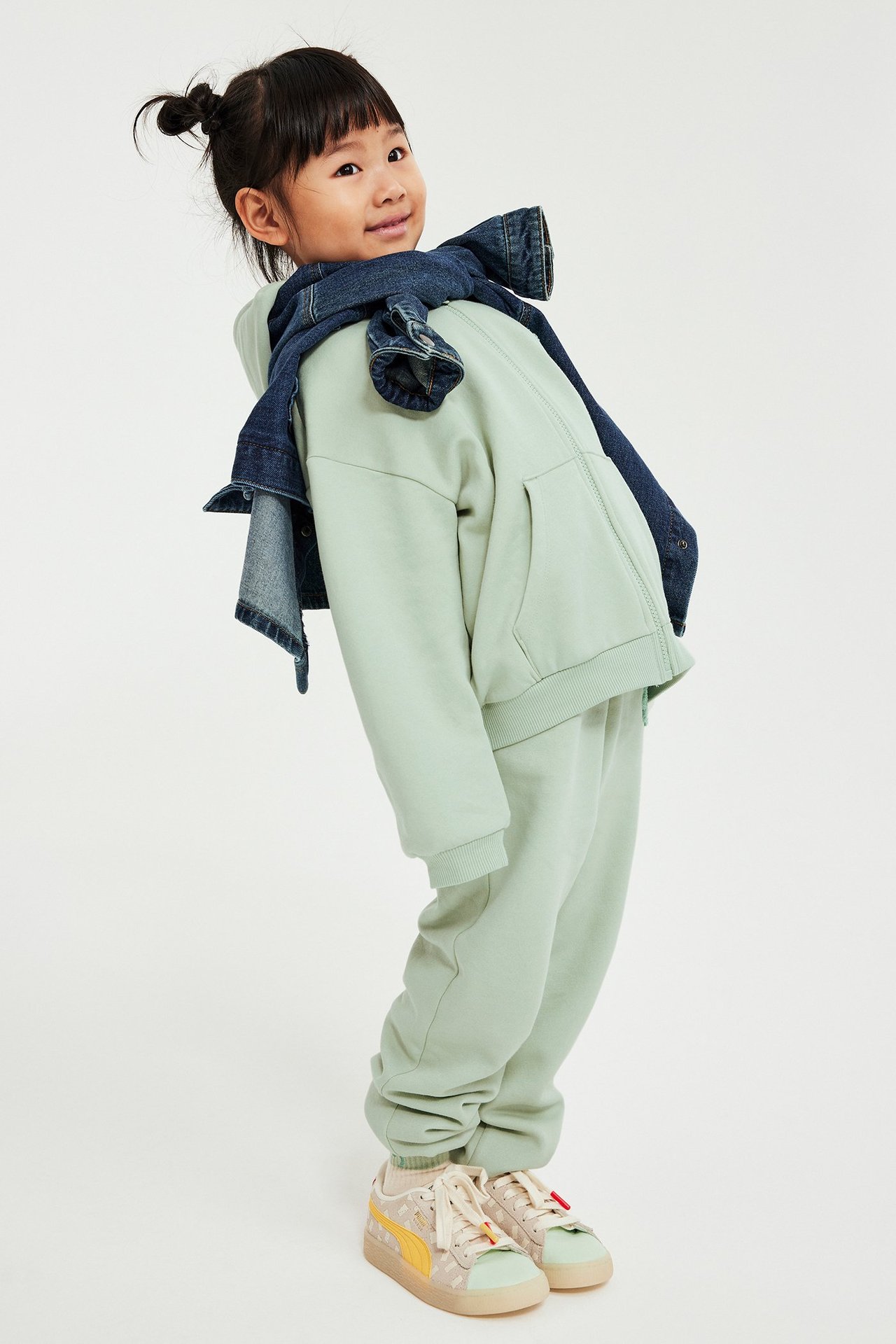 Dziecko w zielonej bluzie na zamek, dresach, niebieska kurtka na ramionach, jasne sneakersy.