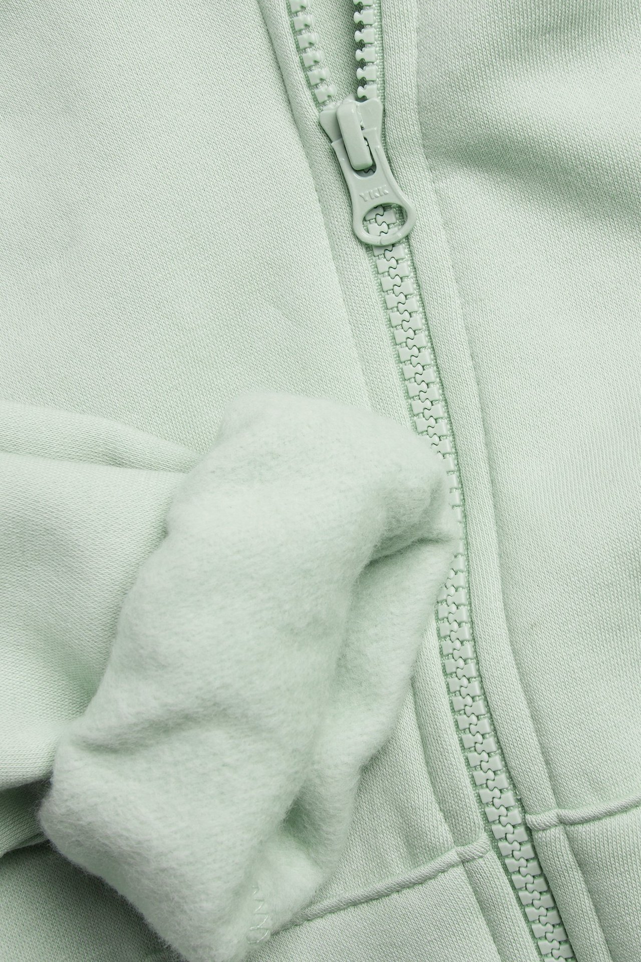 Zbliżenie na jasnozieloną bluzę z kapturem, pokazujące zamek błyskawiczny i jej miękkie, szczotkowane wnętrze.