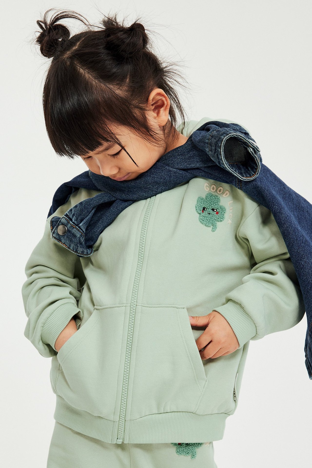 Przód, dziecko w jasnozielonej bluzie na zamek z koniczyną, ciemnoniebieska dżinsowa kurtka narzucona.