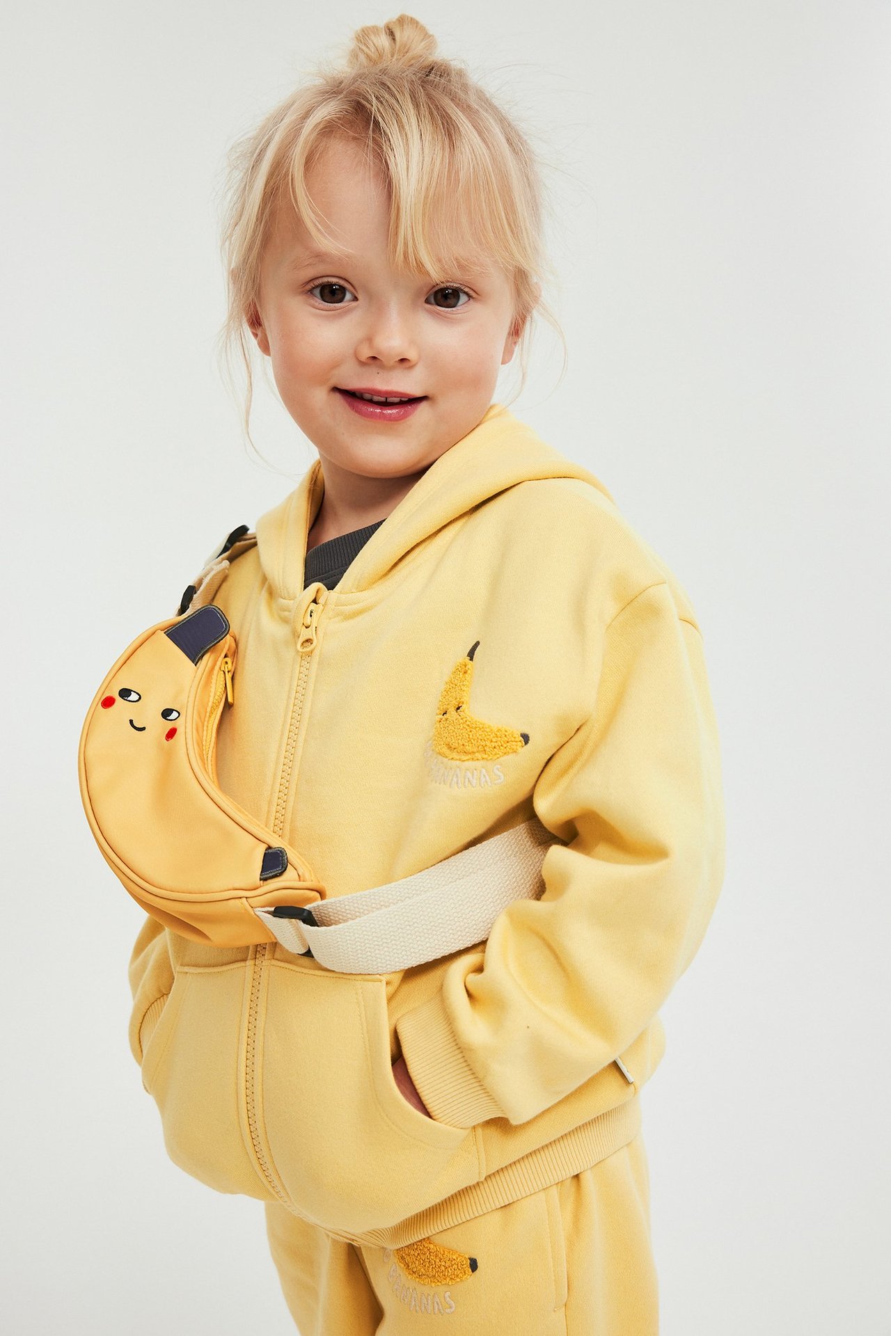 Edestä, lapsi keltaisessa vetoketjuhupparissa, banaanikirjailulla, yhteensopivissa housuissa ja banaanivyölaukussa.