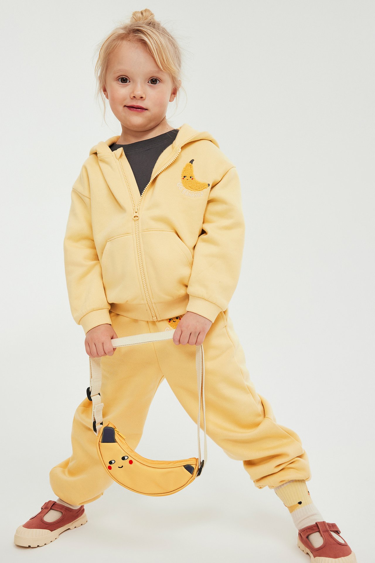 Edestäpäin kuvassa lapsi keltaisessa vetoketjuhupparissa banaanikirjonnalla, keltaisissa collegehousuissa ja banaanilaukkua pidellen.