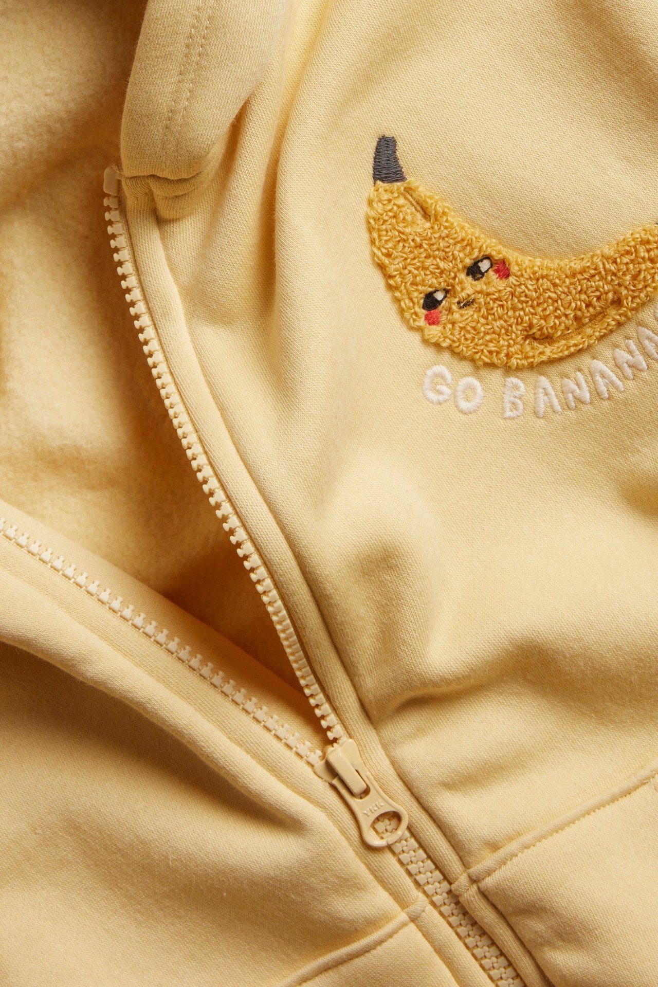 Zbliżenie żółtej rozpinanej bluzy z kapturem z haftem banana i napisem 'GO BANANAS'.