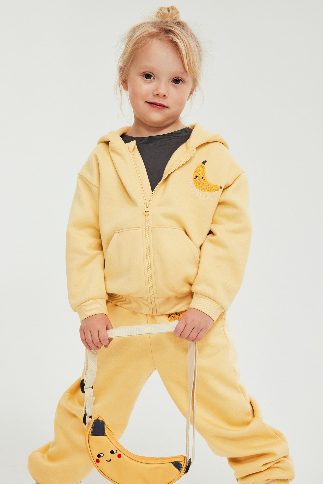 Framifrån: ett barn i gul huvtröja med dragkedja, bananbrodyr, matchande mjukisbyxor och en bananväska.