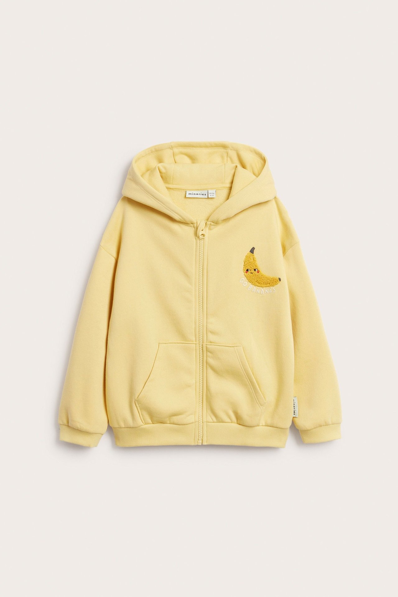 Edestäpäin keltainen lasten huppari puuvillasekoitteesta, vetoketjulla, taskuilla ja brodeeratulla banaanilla.