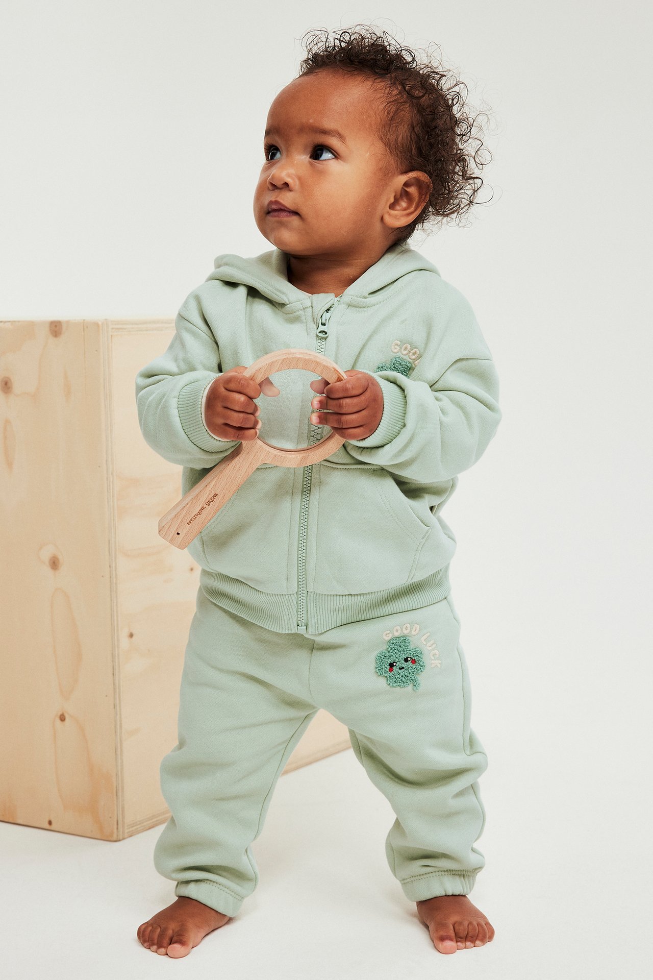 Framifrån: Baby i grön hoodie med dragkedja och klövertryck, matchande mjukisbyxor, håller ett förstoringsglas i trä.