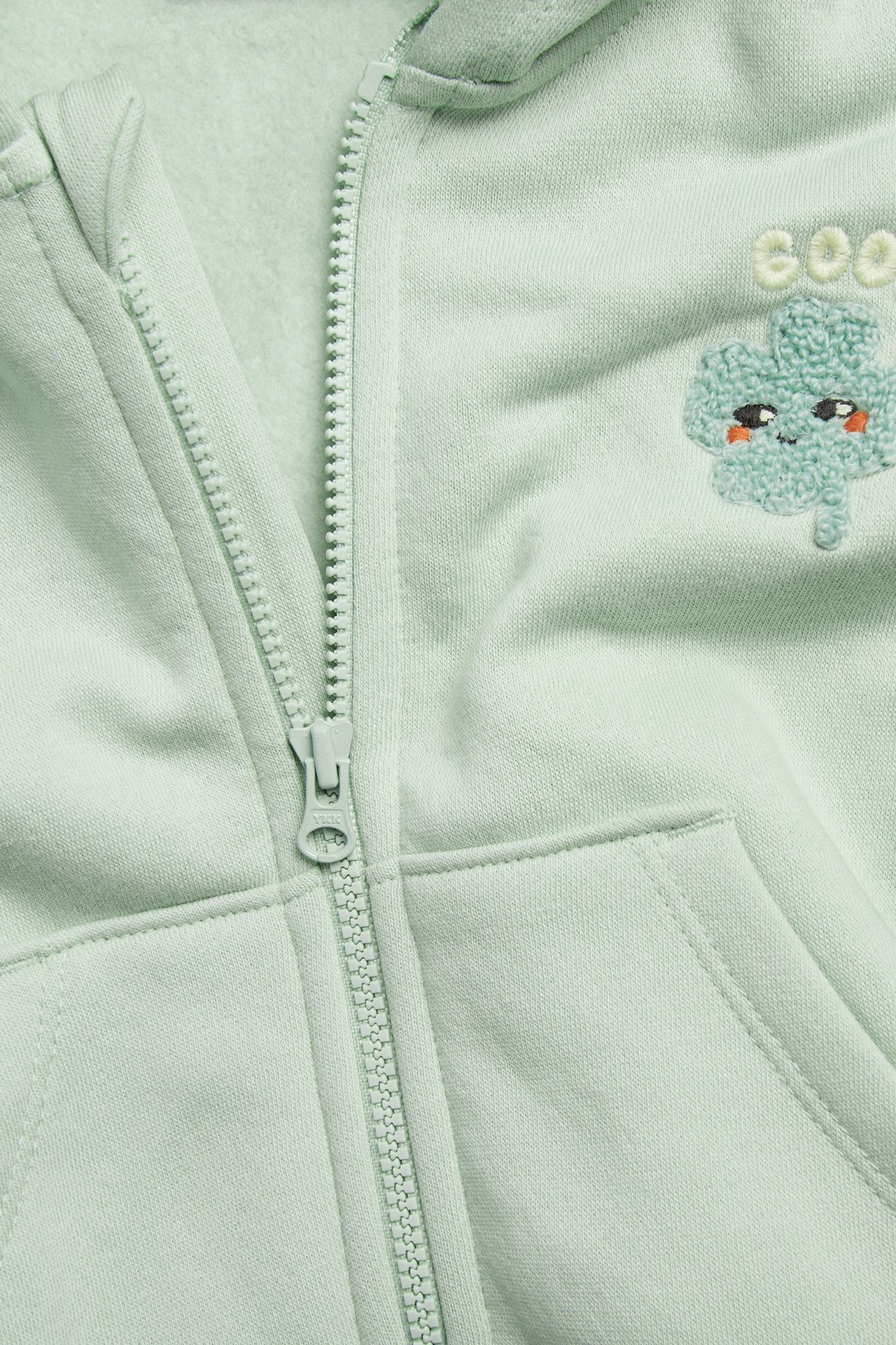 Zbliżenie zielonej bluzy z kapturem, z widocznym zamkiem i haftowaną koniczyną.