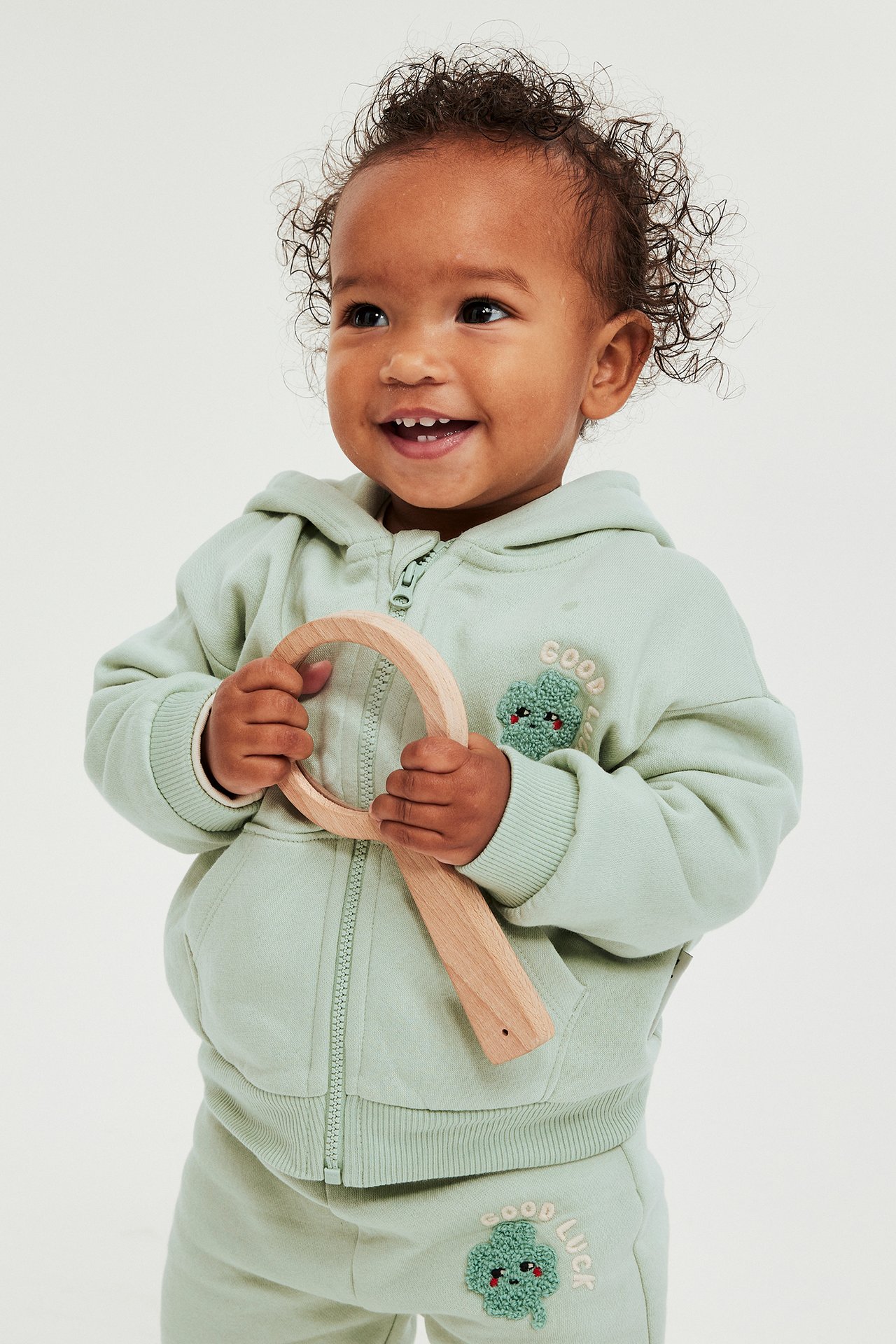 Frontvy av baby i grön hoodie med dragkedja, broderad klöver, matchande mjukisbyxor, håller en träleksak.