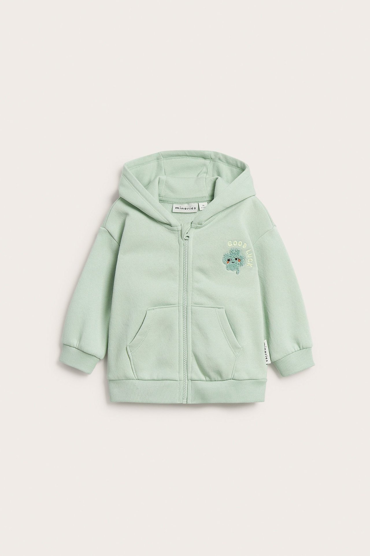 Framifrån: Babyhoodie med dragkedja i ljusgrönt med klöver- och 'Good Luck'-tryck.