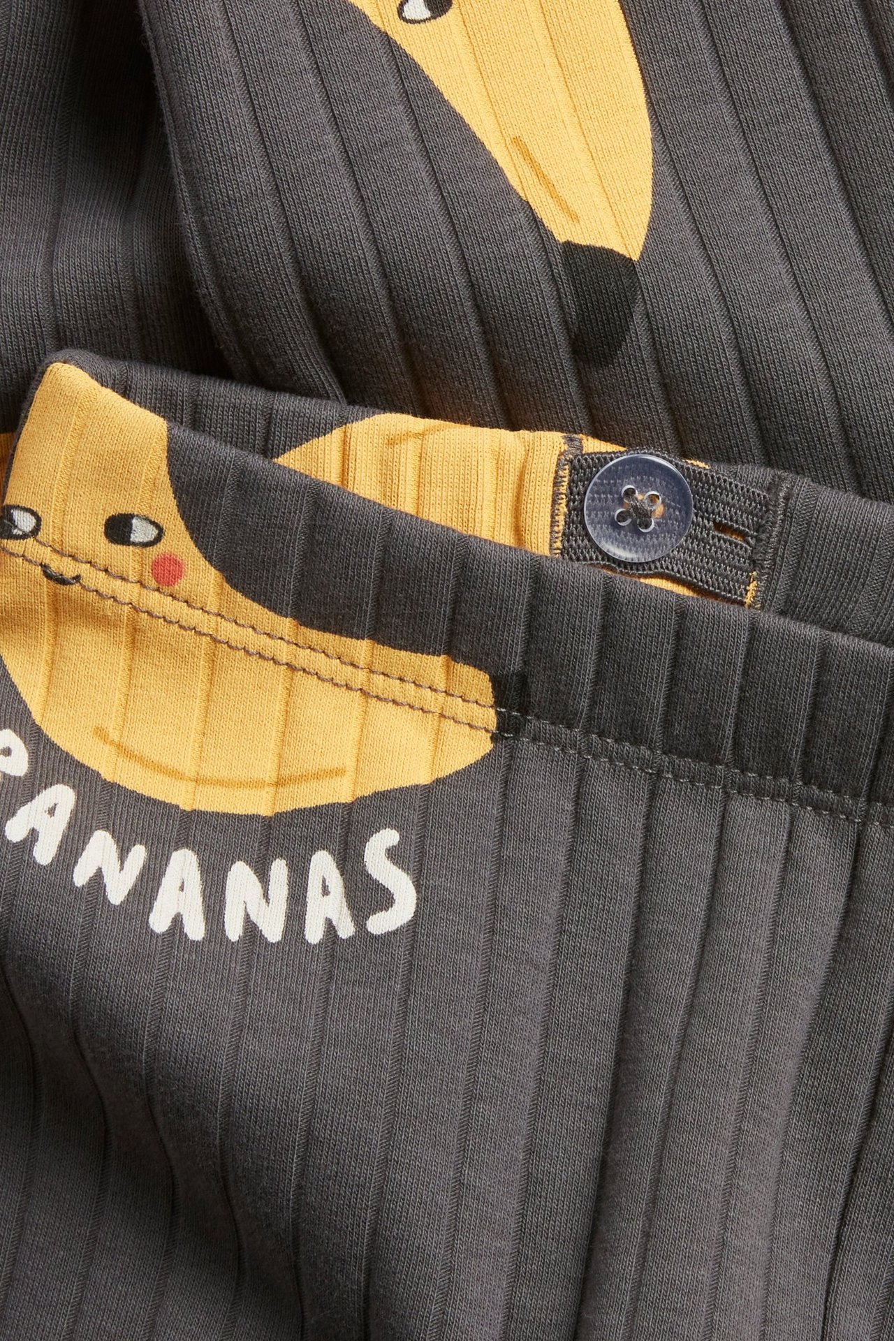 Zbliżenie na ciemnoszare prążkowane legginsy dziecięce z żółtym nadrukiem bananów i regulowanym pasem na guzik.