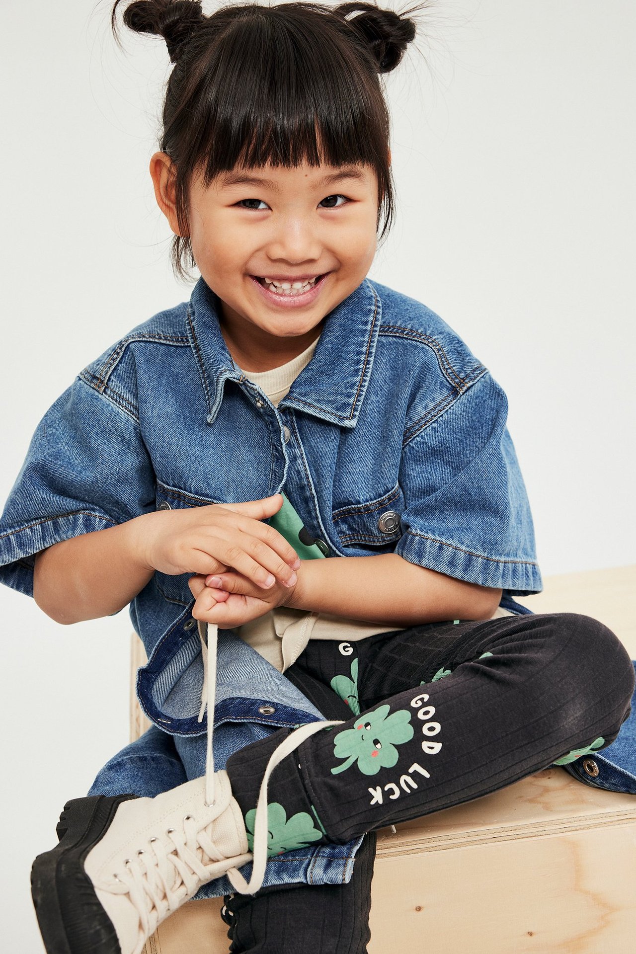 Edestäpäin hymyilevä lapsi istuu, yllään mustat ribbileggingsit vihreällä apilaprintillä ja farkkutakki.