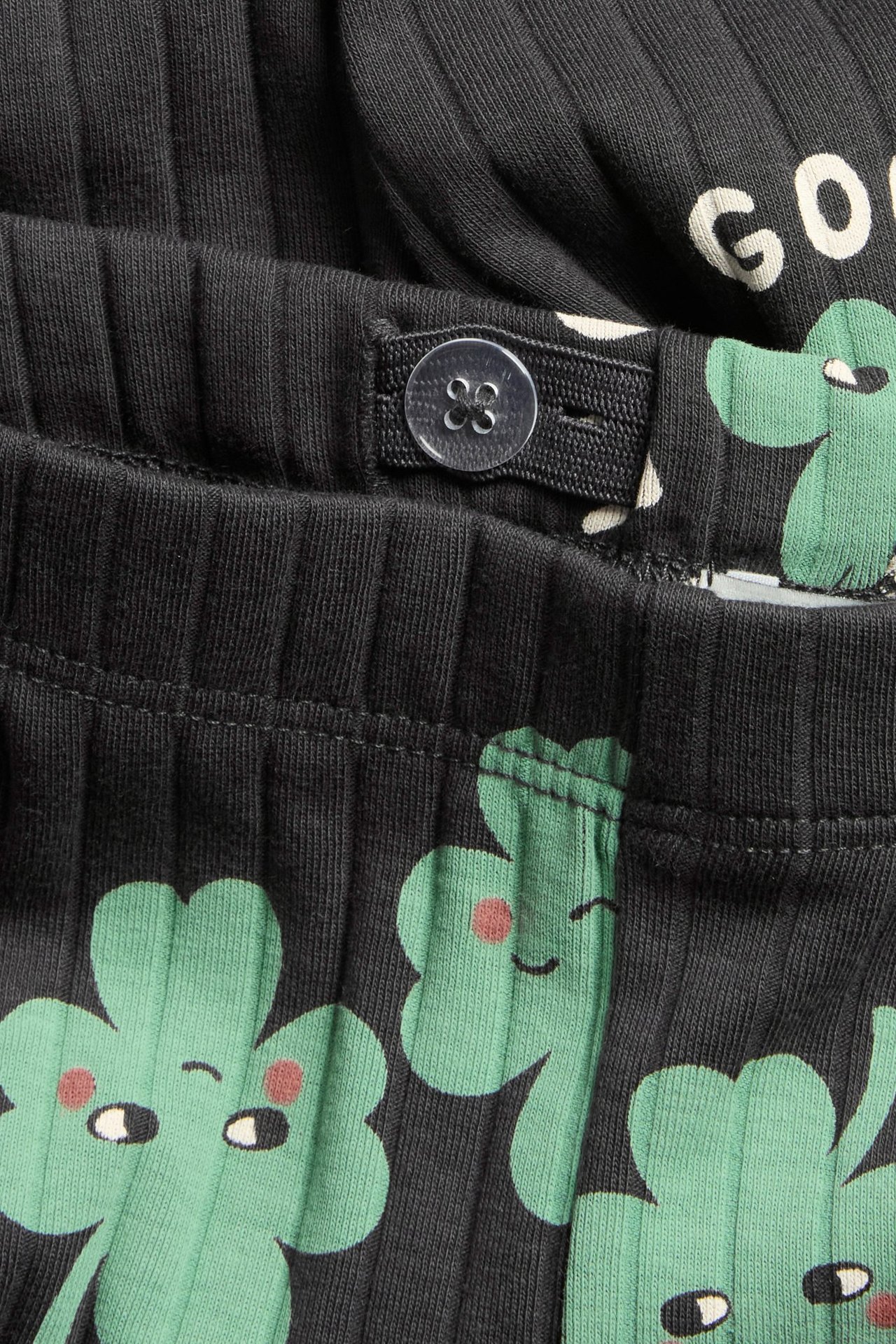 Lähikuva mustista ribatuista lasten leggingseistä, joissa vihreä apilaprintti ja säädettävä vyötärönappi.