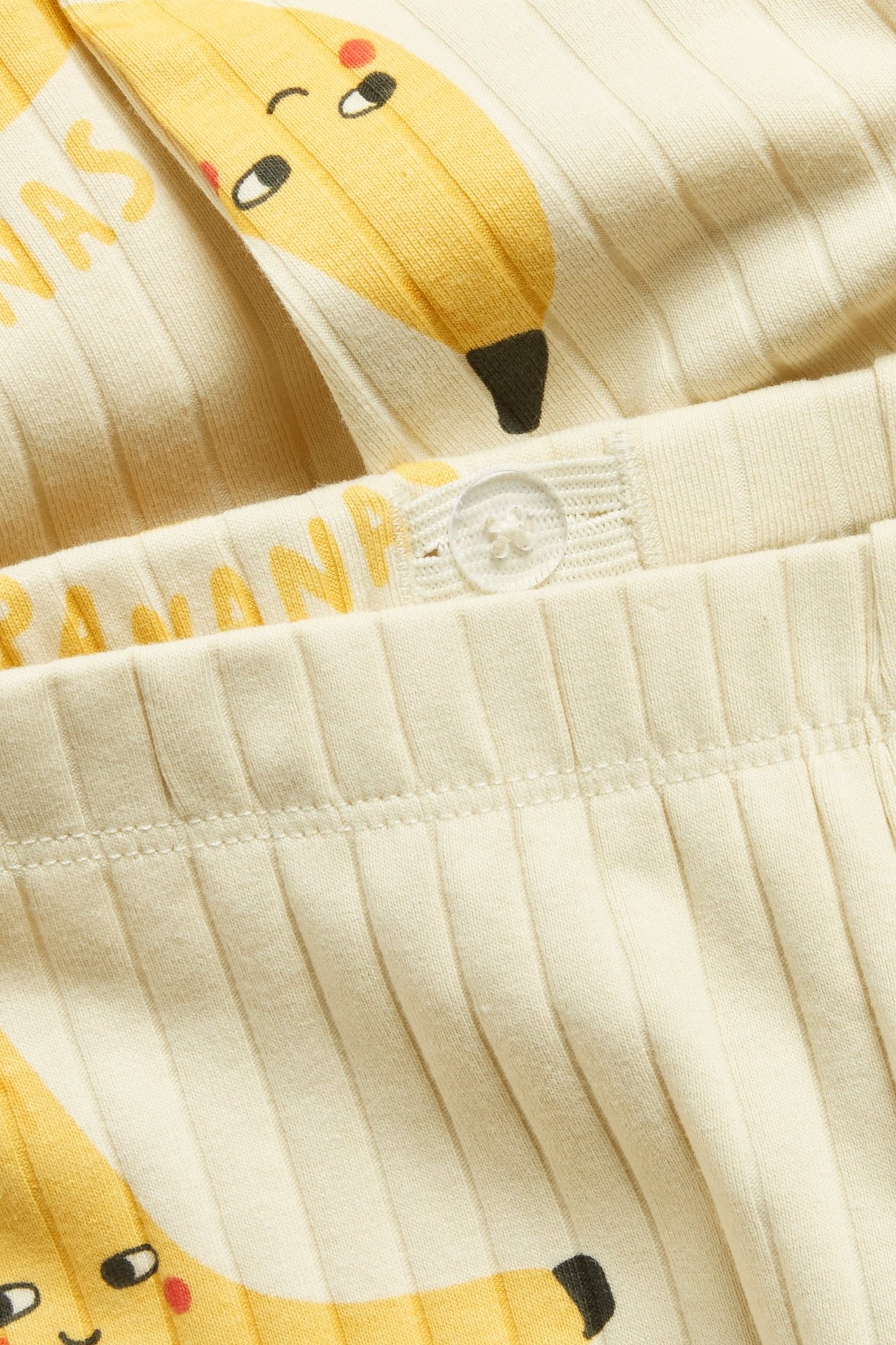 Zbliżenie beżowych prążkowanych legginsów z nadrukiem bananów i regulowaną talią z guzikiem.