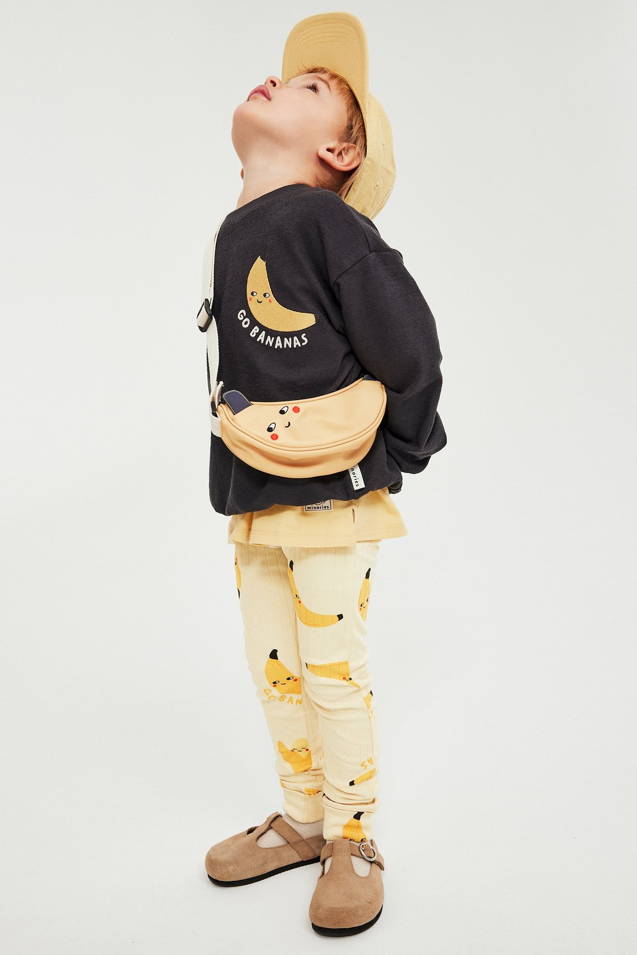 Lapsi yllään beiget ribatut banaanilöysät leggingsit, tummanharmaa collegepaita, keltainen toppi, beige lippis, banaanivyölaukku.
