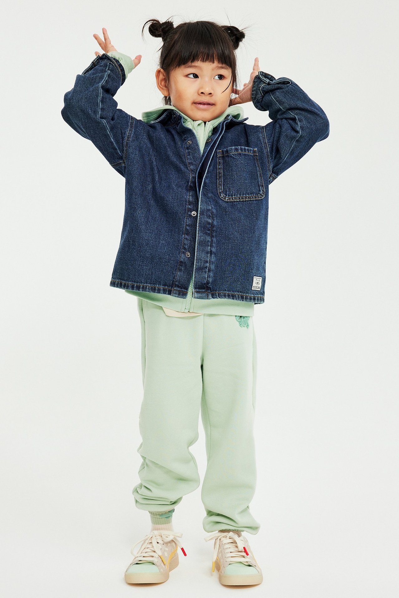 Barn i grønne joggebukser med applikasjon, lys grønn hoodie og blå dongeriskjorte, sett forfra.