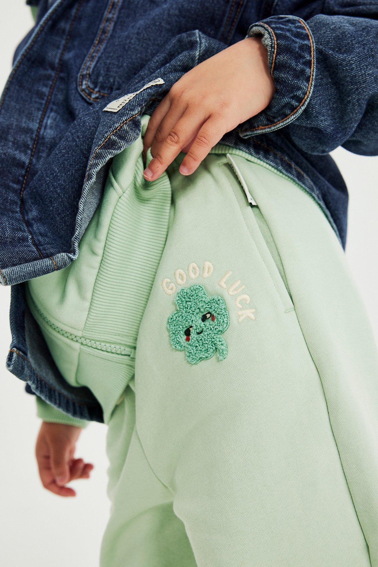 Zbliżenie dziecka w jasnozielonych spodniach dresowych z aplikacją koniczyny i granatowej kurtce dżinsowej.