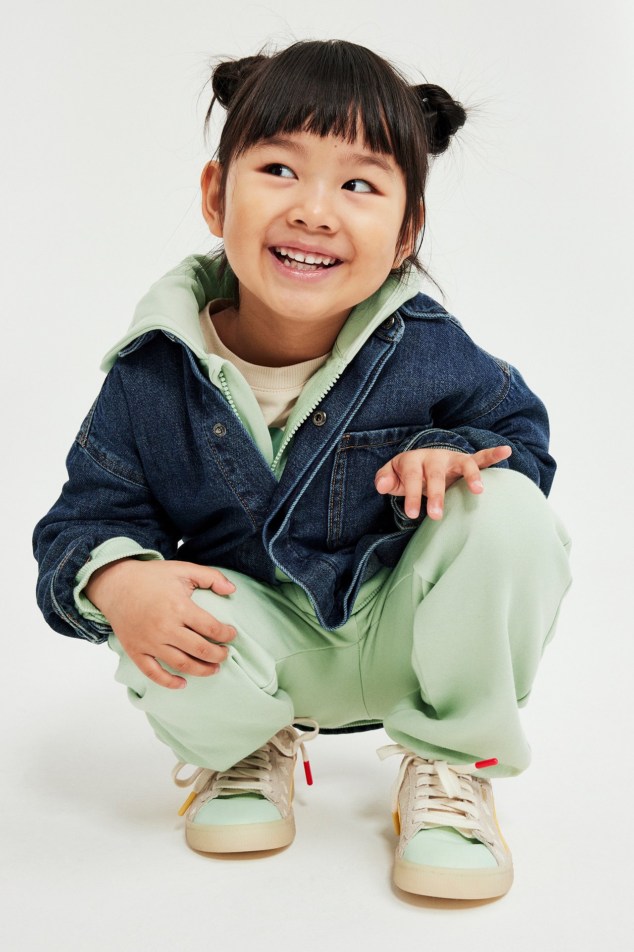 Widok z przodu: Dziecko w zielonych spodniach dresowych, jasnozielonej bluzie, beżowym t-shircie i niebieskiej kurtce dżinsowej.