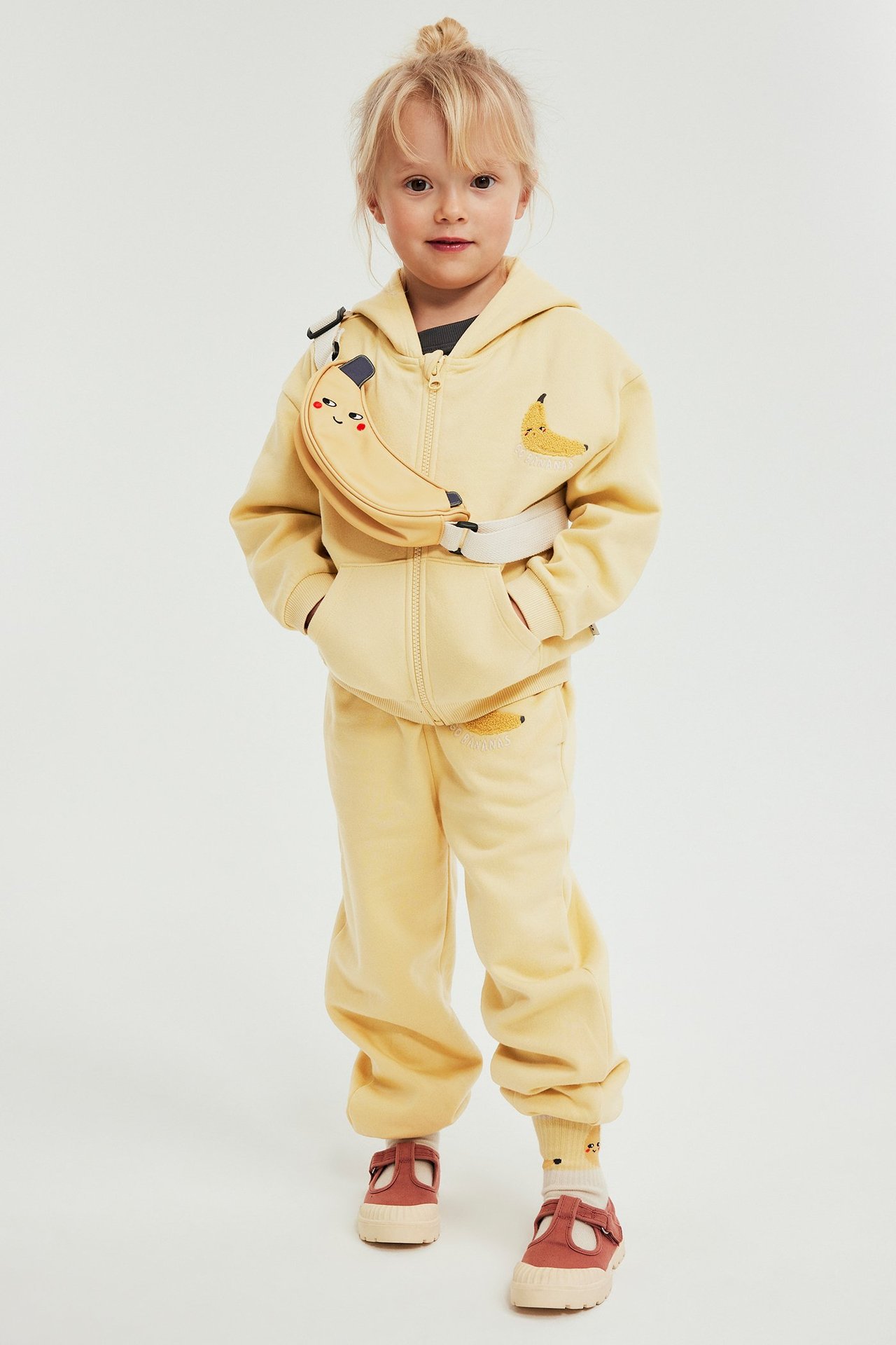Edestäpäin: Lapsi keltaisissa banaanikuvioisissa collegehousuissa, yhteensopivassa hupparissa ja banaanivyölaukussa.