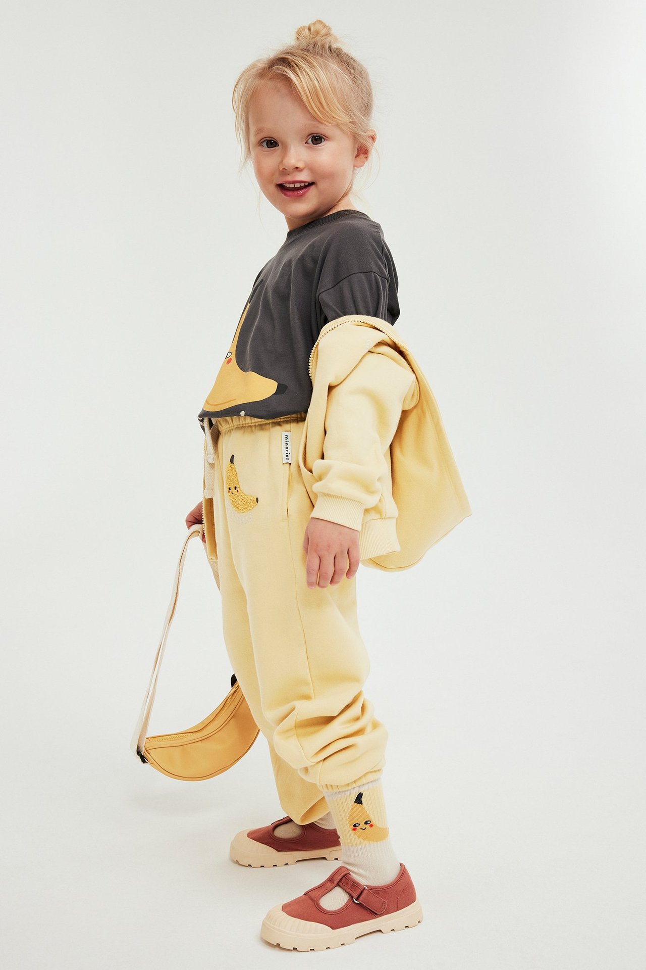 Barn bär gula mjukisbyxor med bananapplikation, grå t-shirt, gul huvtröja, bananstrumpor, bruna skor, bananväska.