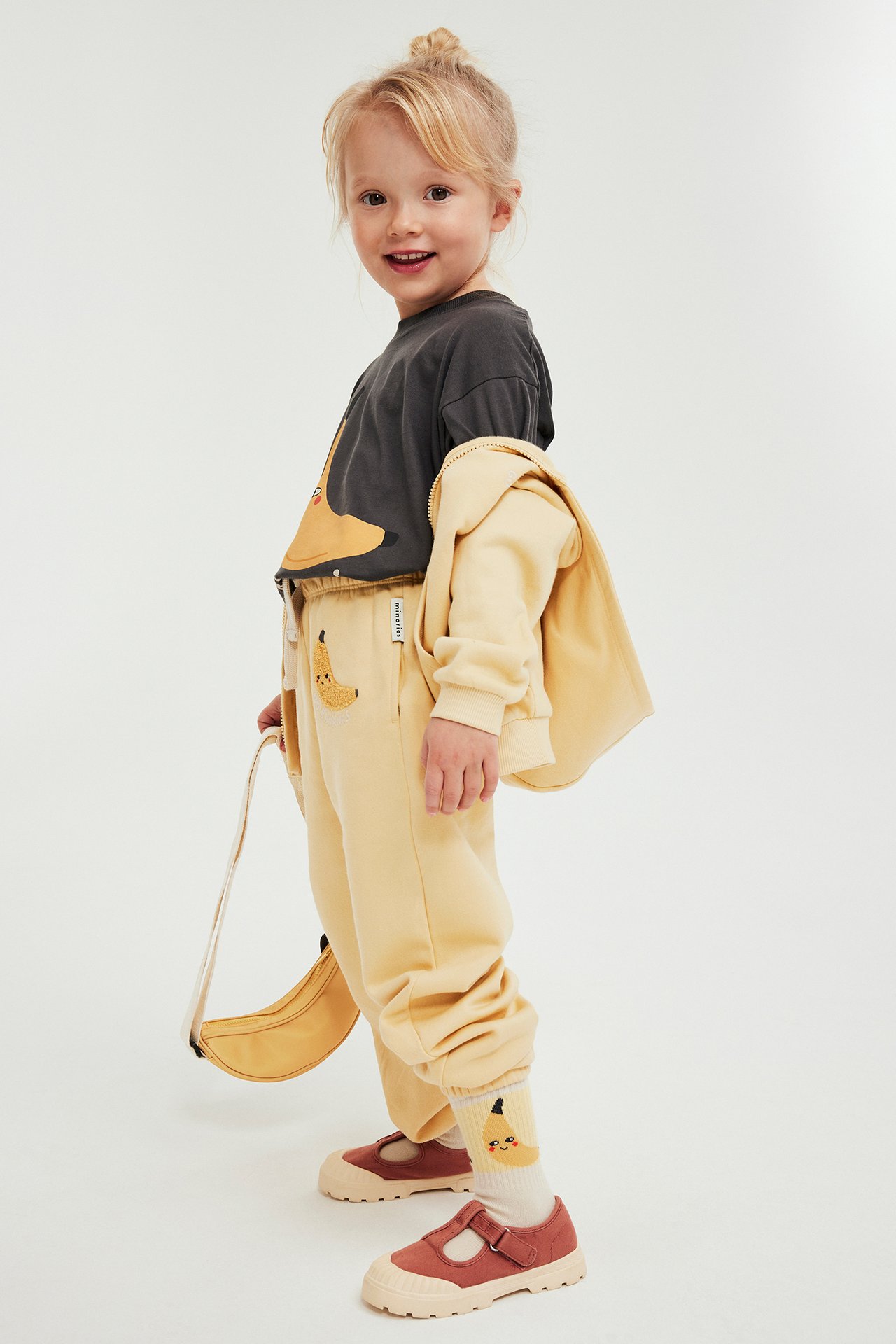 Barn bär gula mjukisbyxor med bananapplikation, mörkgrå t-shirt, gul huvtröja, bananstrumpor och rödbruna skor.