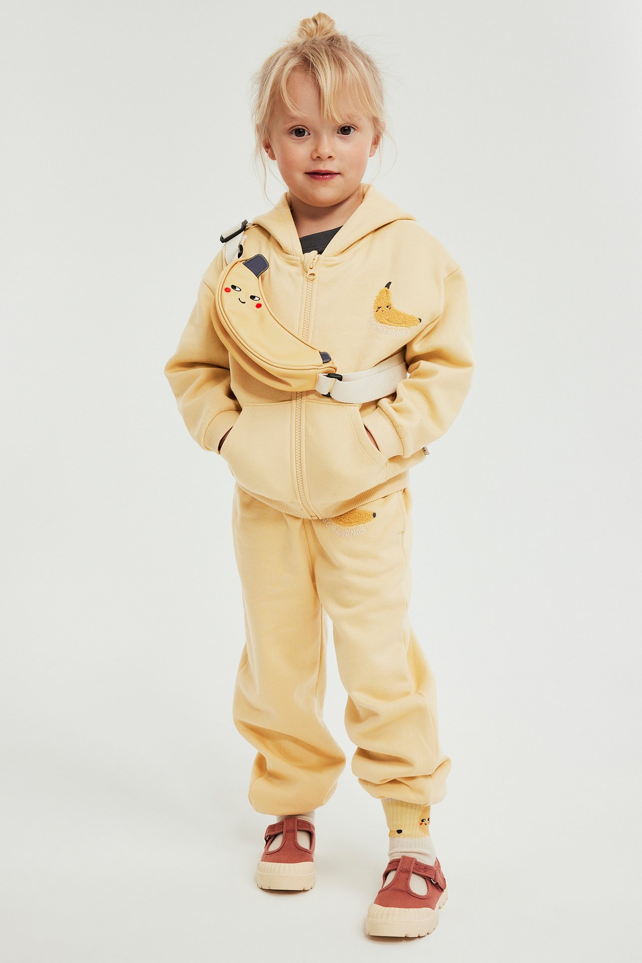 Lapsi yllään keltaiset collegehousut banaanikirjonnalla, yhteensopiva vetoketjuhuppari, banaani-vyölaukku ja ruosteenpunaiset kengät.