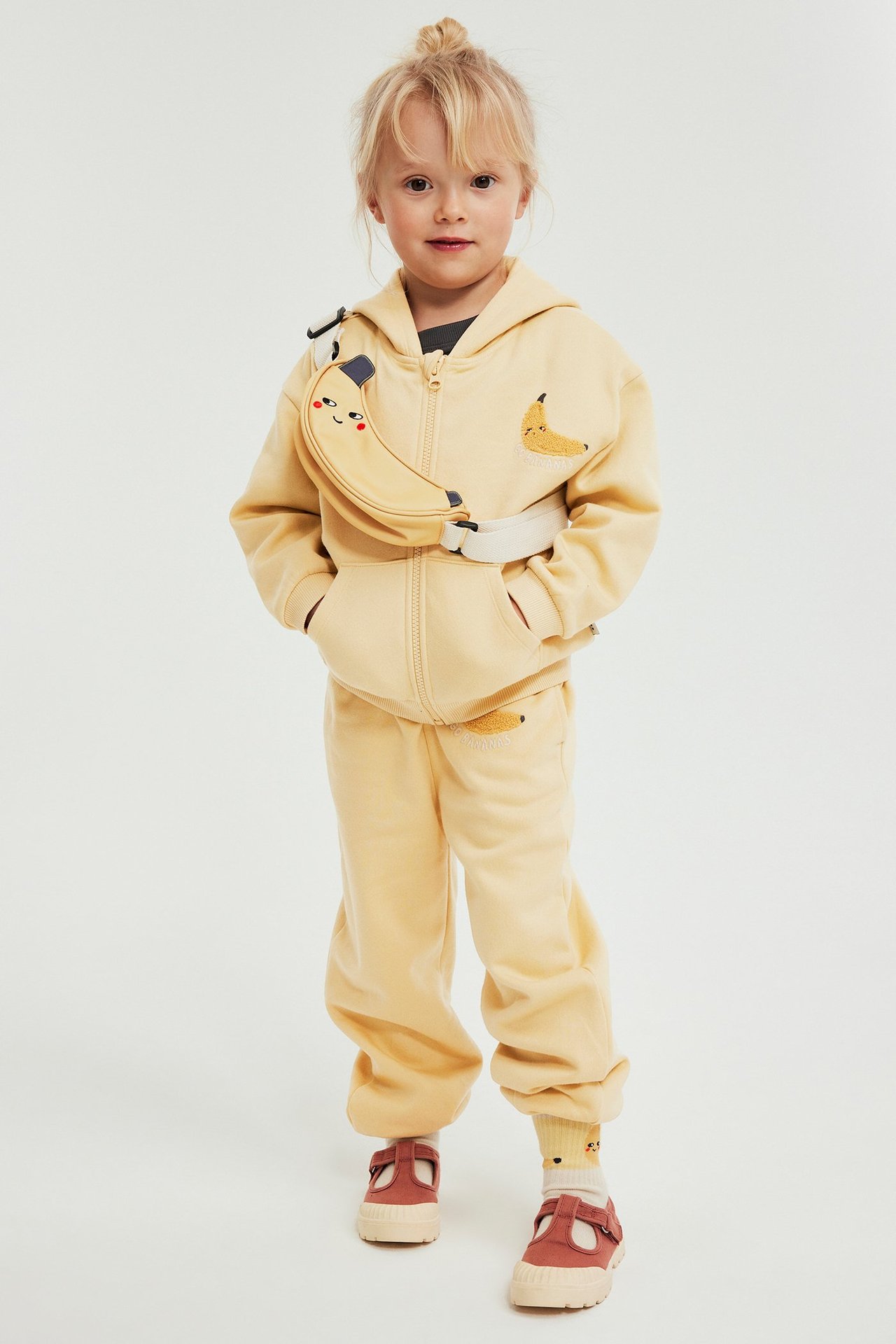 Lapsi yllään keltaiset collegehousut banaanikirjonnalla, yhteensopiva vetoketjuhuppari, banaani-vyölaukku ja ruosteenpunaiset kengät.