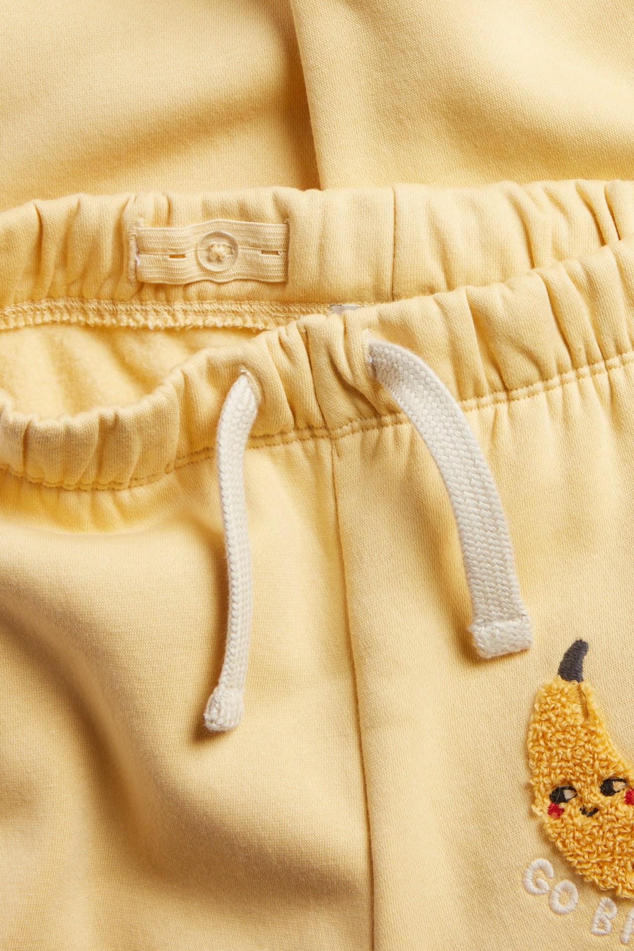 Zbliżenie na żółte dresowe spodnie dziecięce z elastycznym pasem, regulowanym guzikiem, sznurkiem i aplikacją banana.