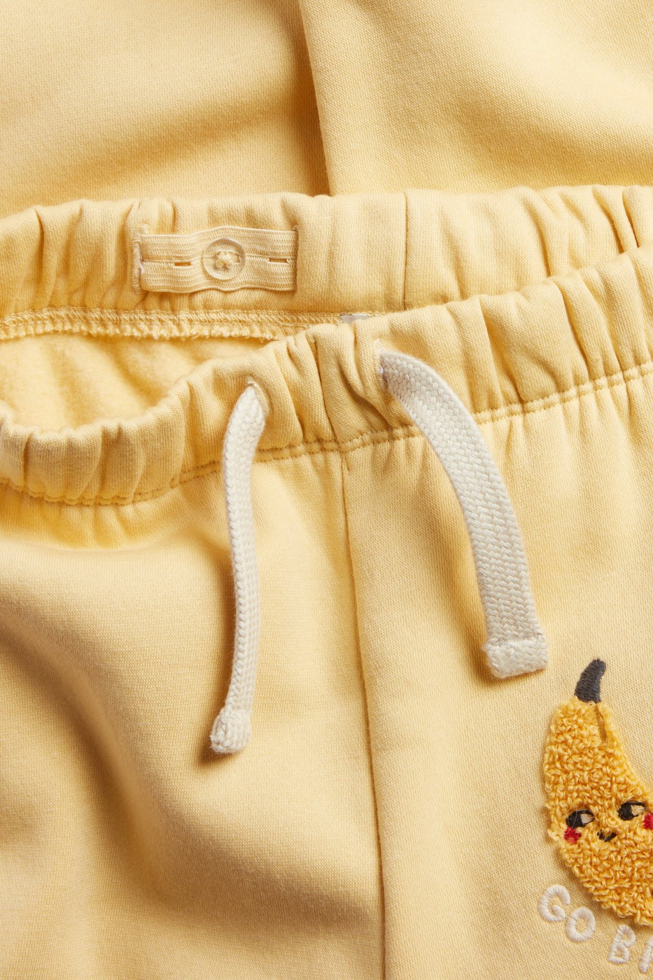 Zbliżenie na żółte dresowe spodnie dziecięce z elastycznym pasem, regulowanym guzikiem, sznurkiem i aplikacją banana.