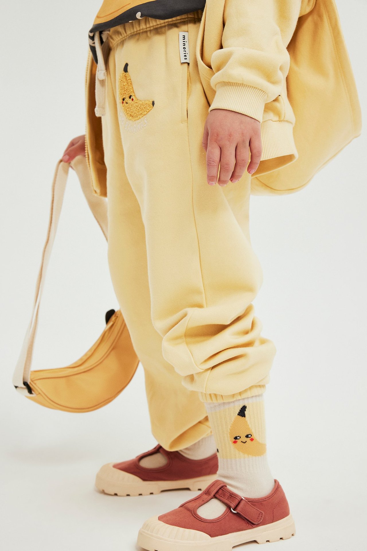 Lapsi keltaisissa collegehousuissa, joissa banaaniapplikaatio, keltainen pusero, kuviolliset sukat, ruskeat kengät ja banaanilaukku.