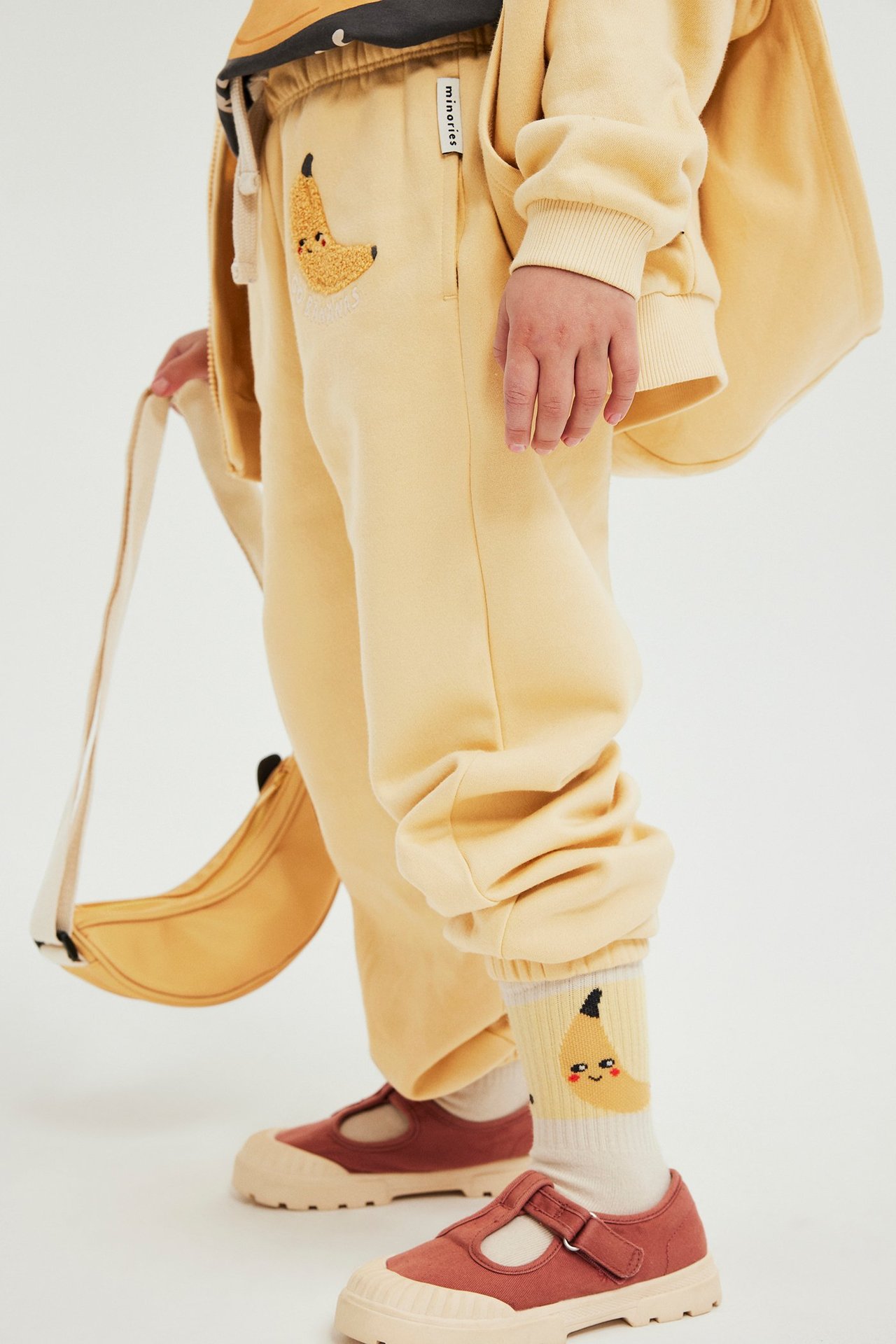 Gule joggebukser til barn med bananapplikasjon, matchende hettegenser, banansokker, rustfargede sko og en bananveske.