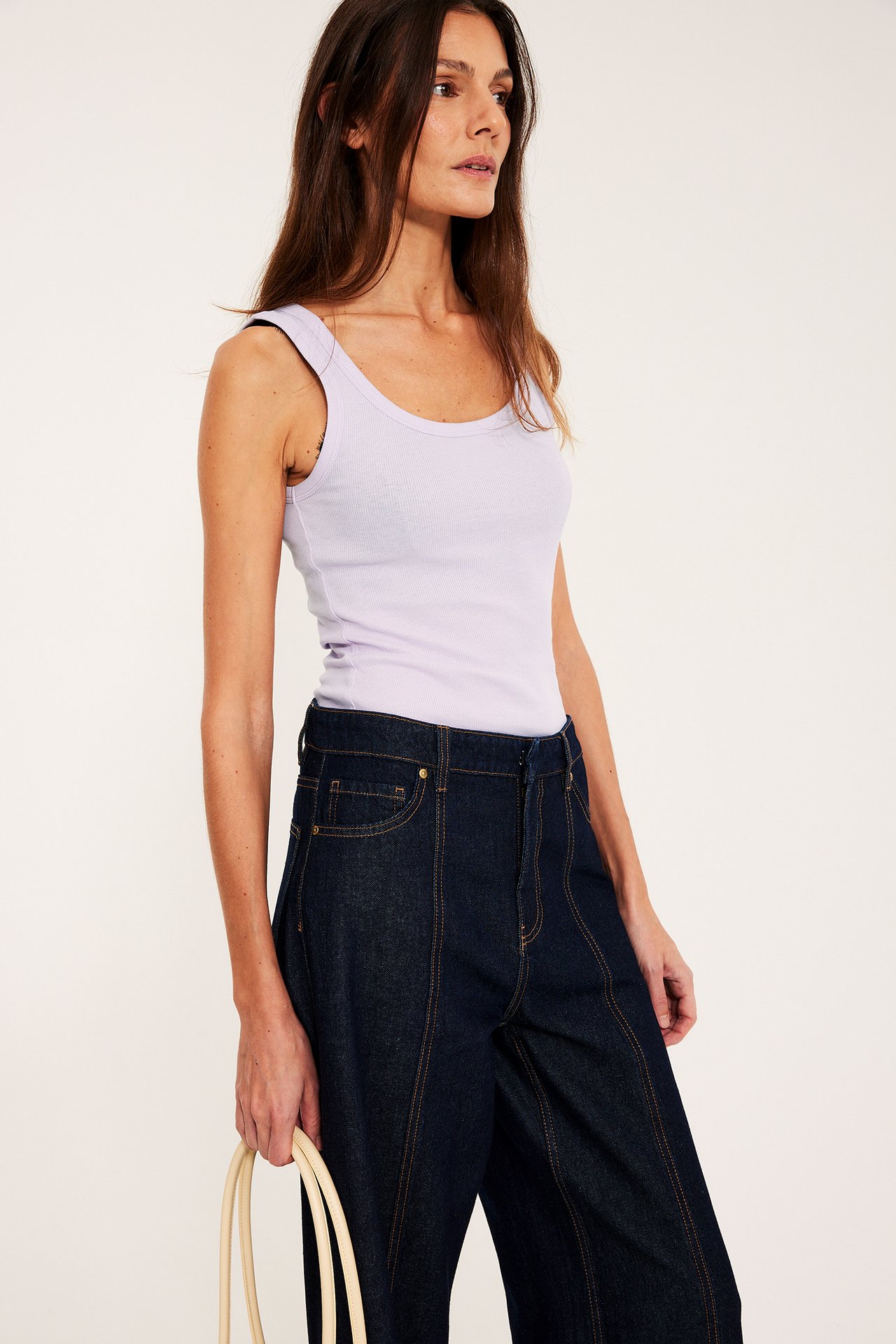 Etunäkymä: nainen yllään vaalea lila ribattu toppi, tummat leveälahkeiset farkut, pidellen vaaleaa laukkua.