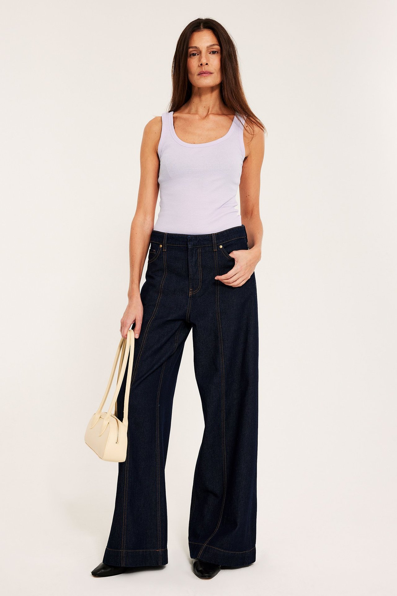 Edestä: Nainen yllään vaaleanlila ribattu toppi, tummat leveälahkeiset farkut ja vaalea käsilaukku.