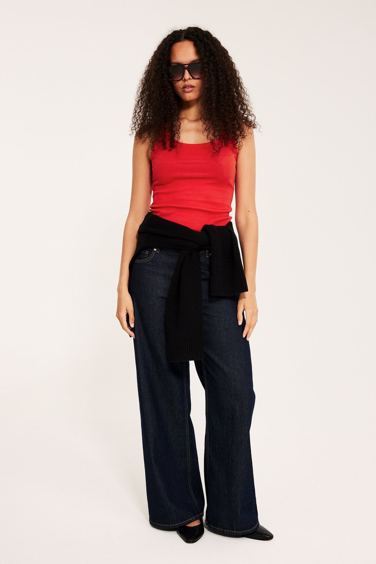 Edestä: Nainen punaisessa ribbitoppissa, tummansinisissä farkuissa, musta pusero vyötäröllä, aurinkolasit.