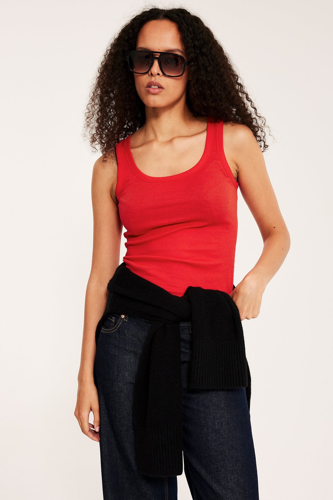 Nainen edestä: punainen ribbitoppi, tummansiniset farkut, musta neule vyötäröllä ja tummat aurinkolasit.