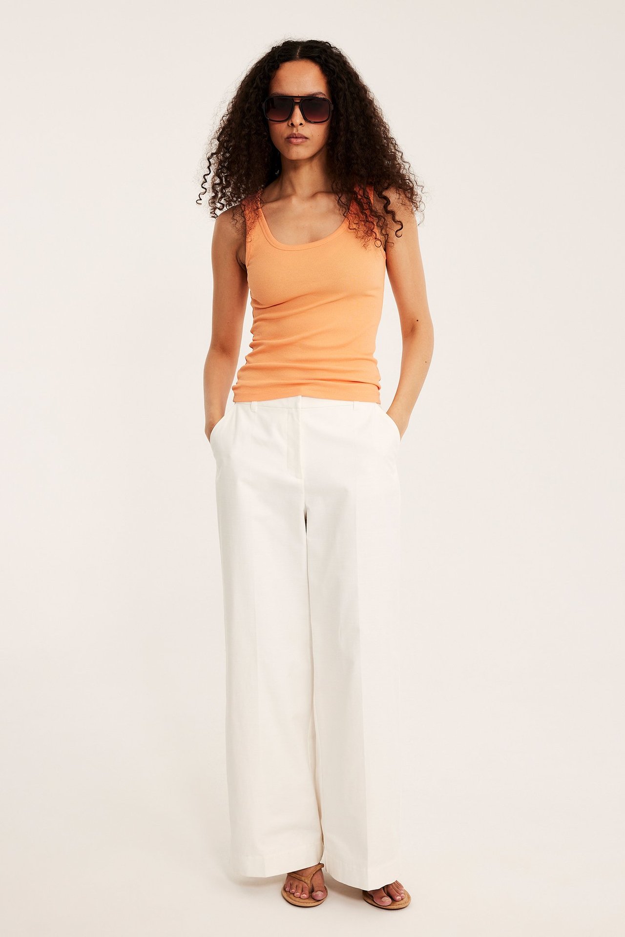 Framifrån, kvinna i orange ribbat linne, vita vida byxor, mörka solglasögon och bruna sandaler.