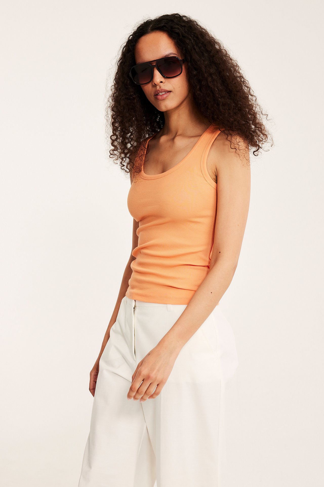 Framifrån: kvinna i orange ribbat linne, vita vida byxor och mörka solglasögon.