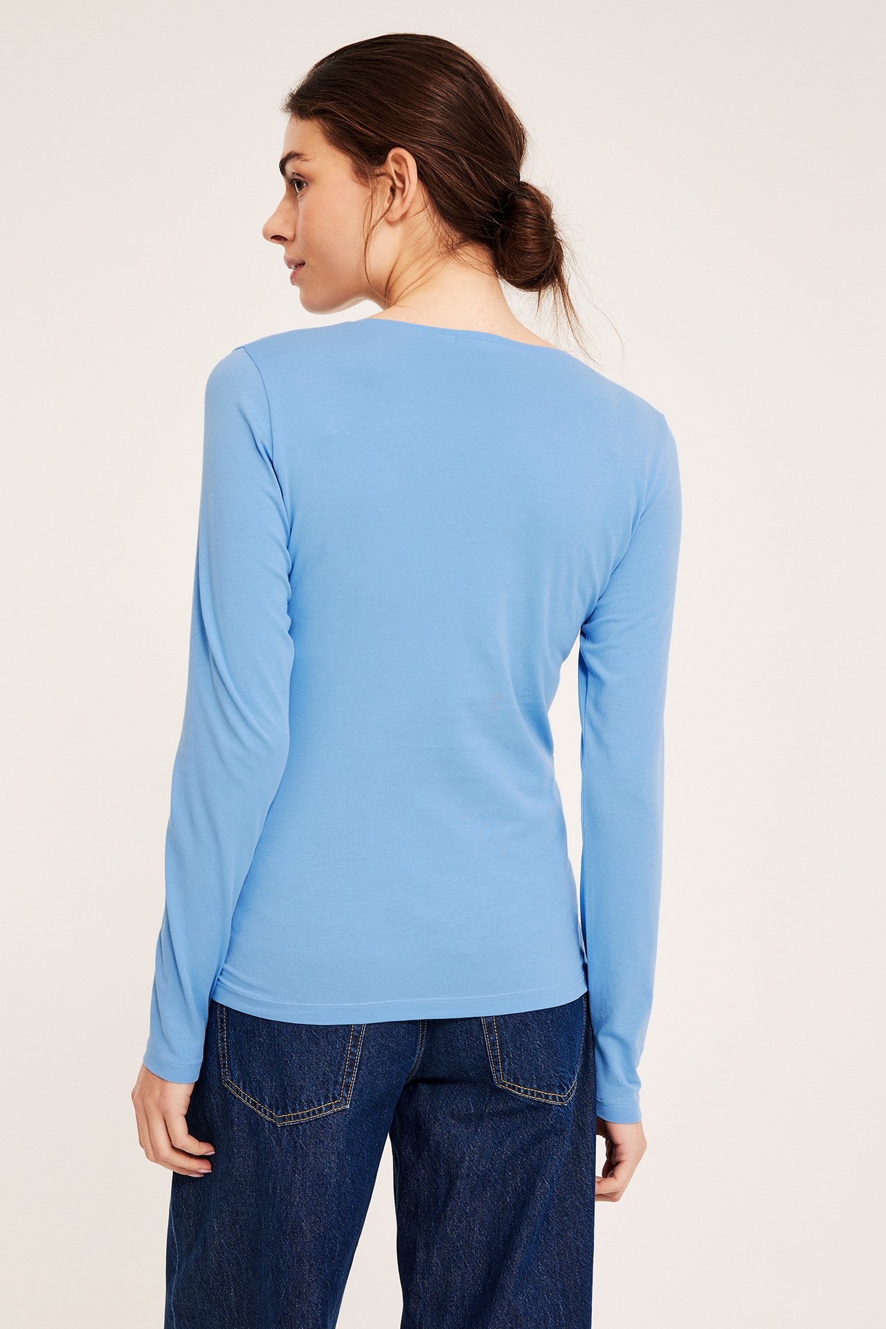 Bakfra: kvinne i lys blå, figurnær, langermet basetopp og mørkeblå jeans.