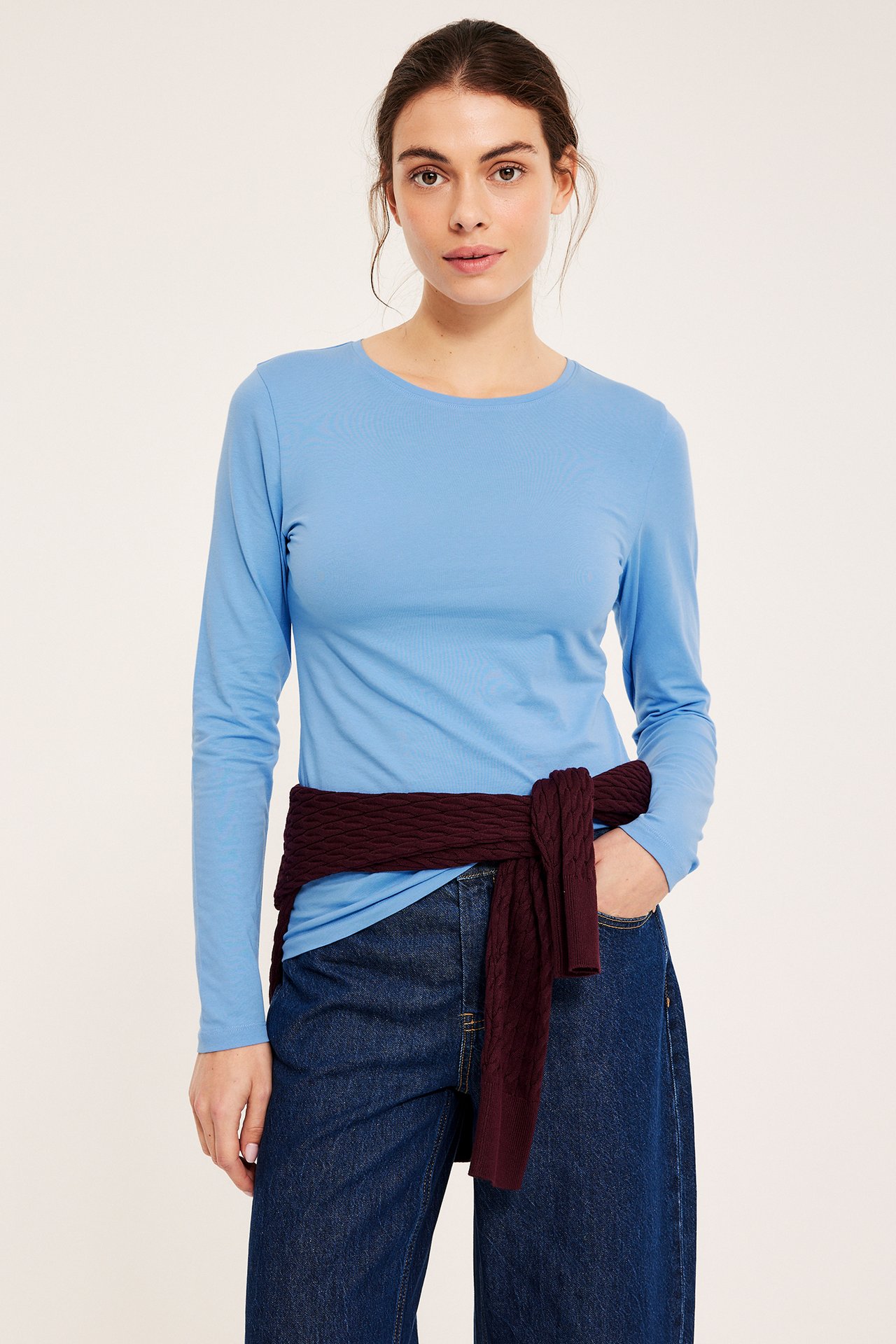 Kvinne i lyseblå langermet topp, mørkeblå jeans og burgunder genser knyttet i midjen.