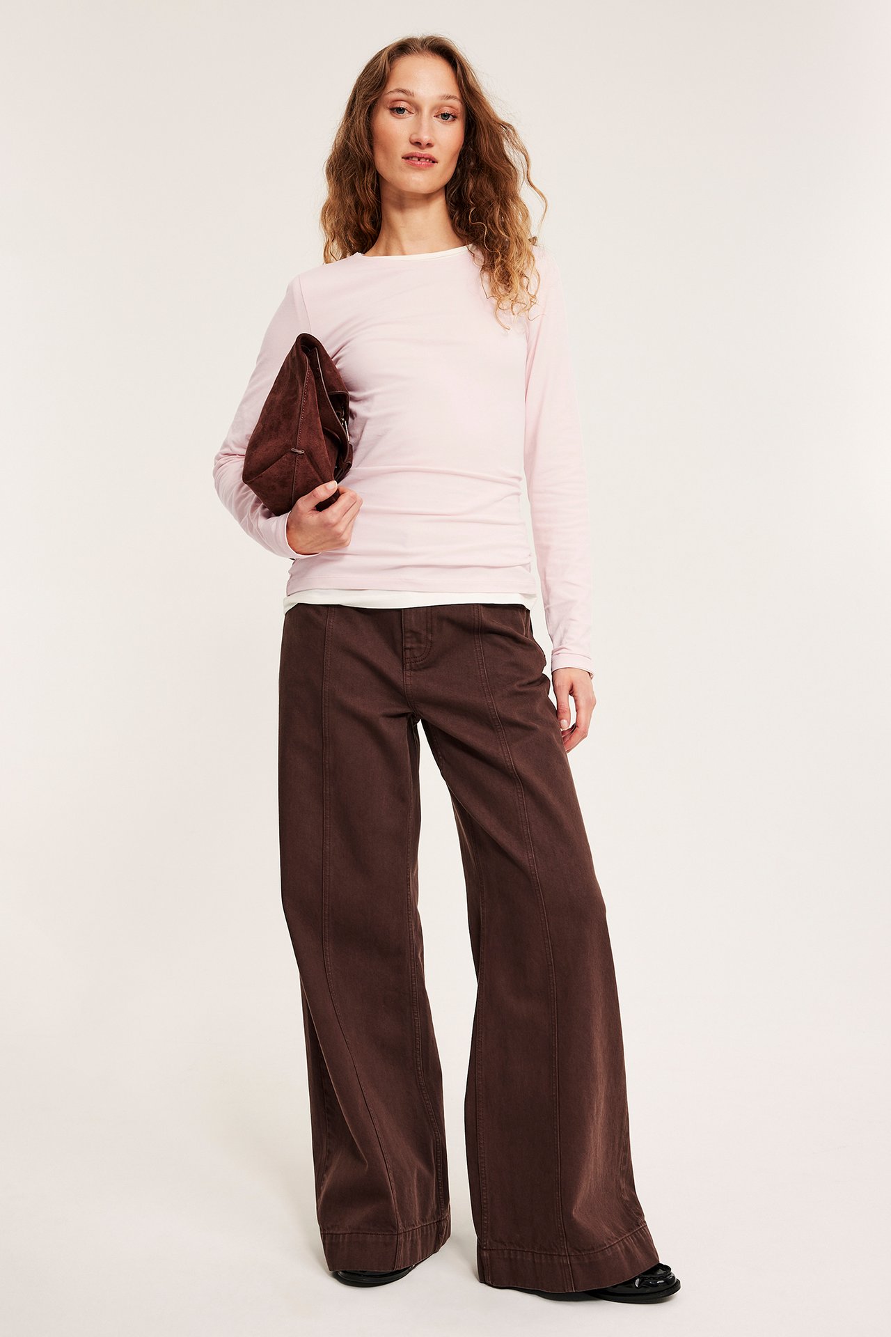 Forfra, kvinne i en tettsittende lys rosa langermet basistopp, brune vide bukser, holder en mørkbrun veske.