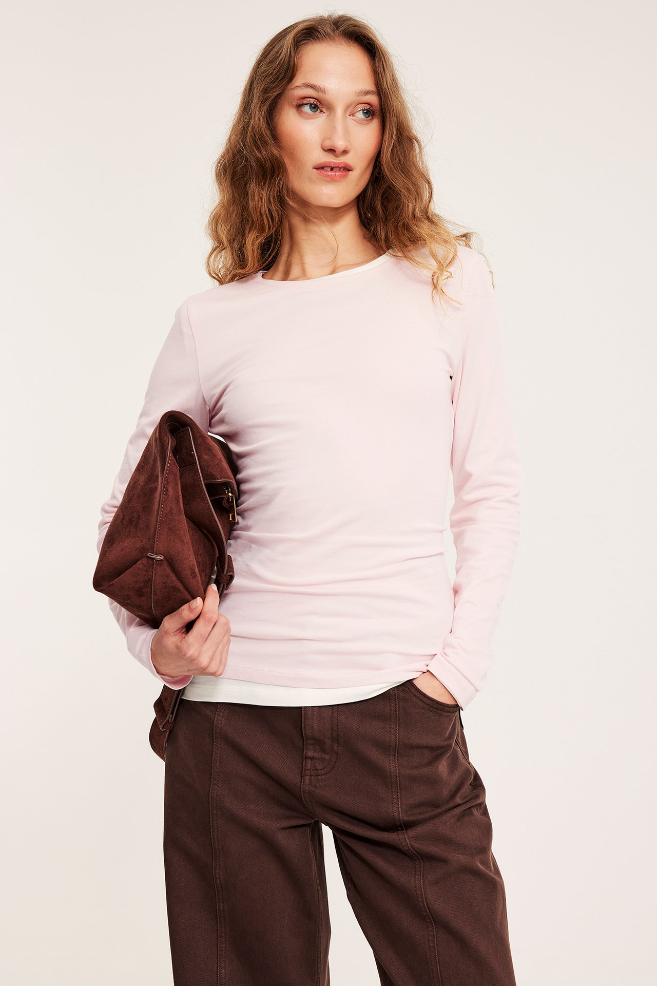 Forfra: en kvinne i lys rosa langermet basistopp, brune bukser, og holder en brun clutchveske.